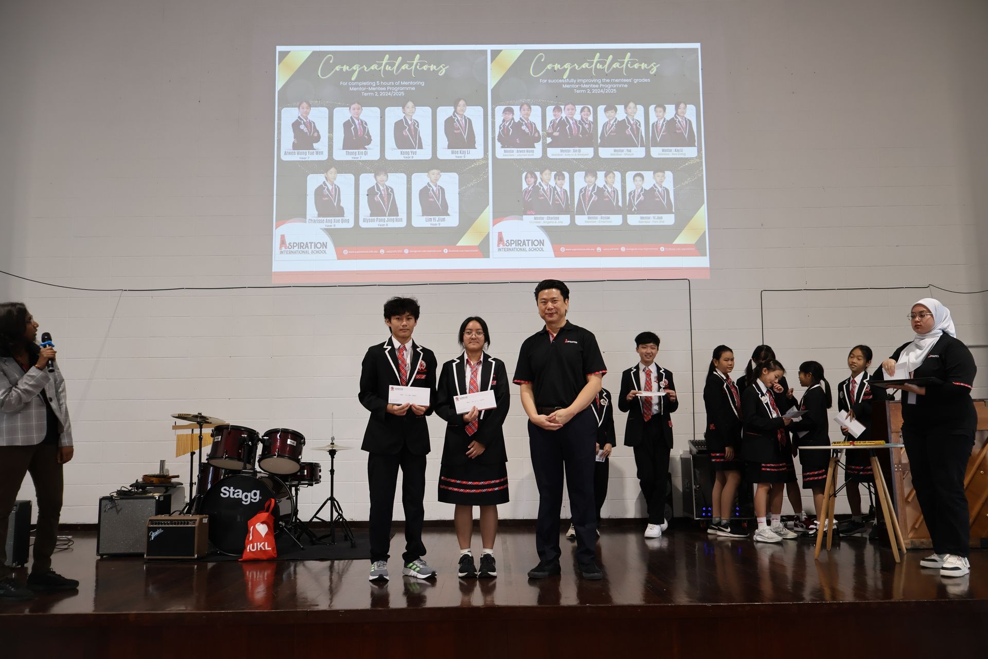 Students on stage with certificates, receiving an award in front of a projector screen with graduation photos.