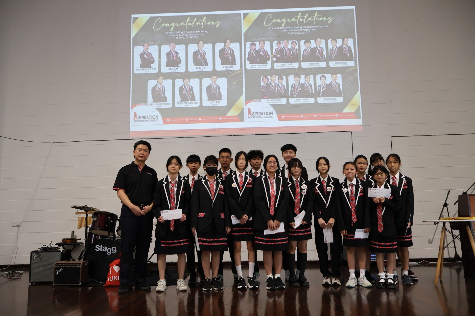 A group of students in uniforms with a teacher on a stage, holding certificates below a congratulatory banner.