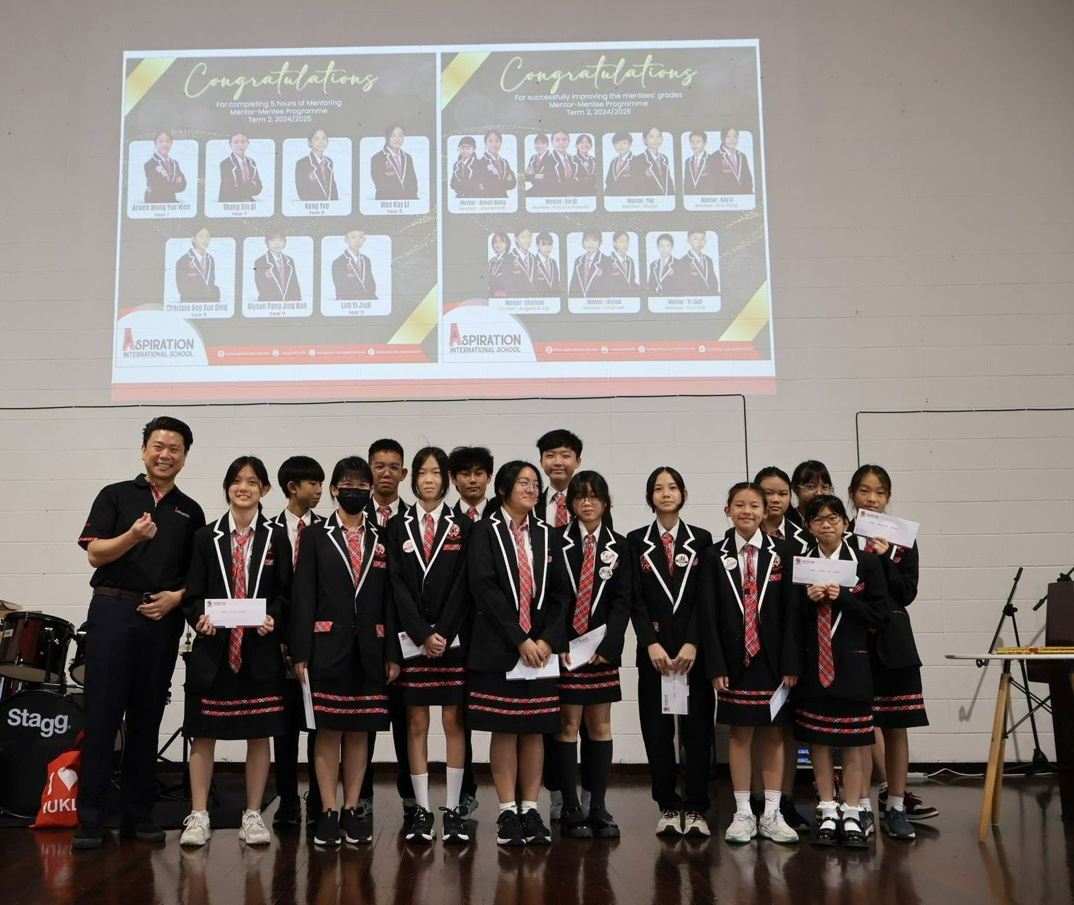 Group of students in uniforms holding certificates posing with a man in front of a projector screen with 