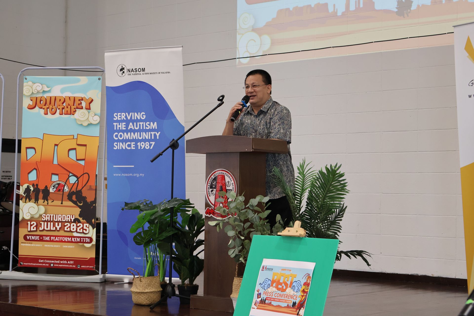Man speaking at a podium, event stage with banners and plants.