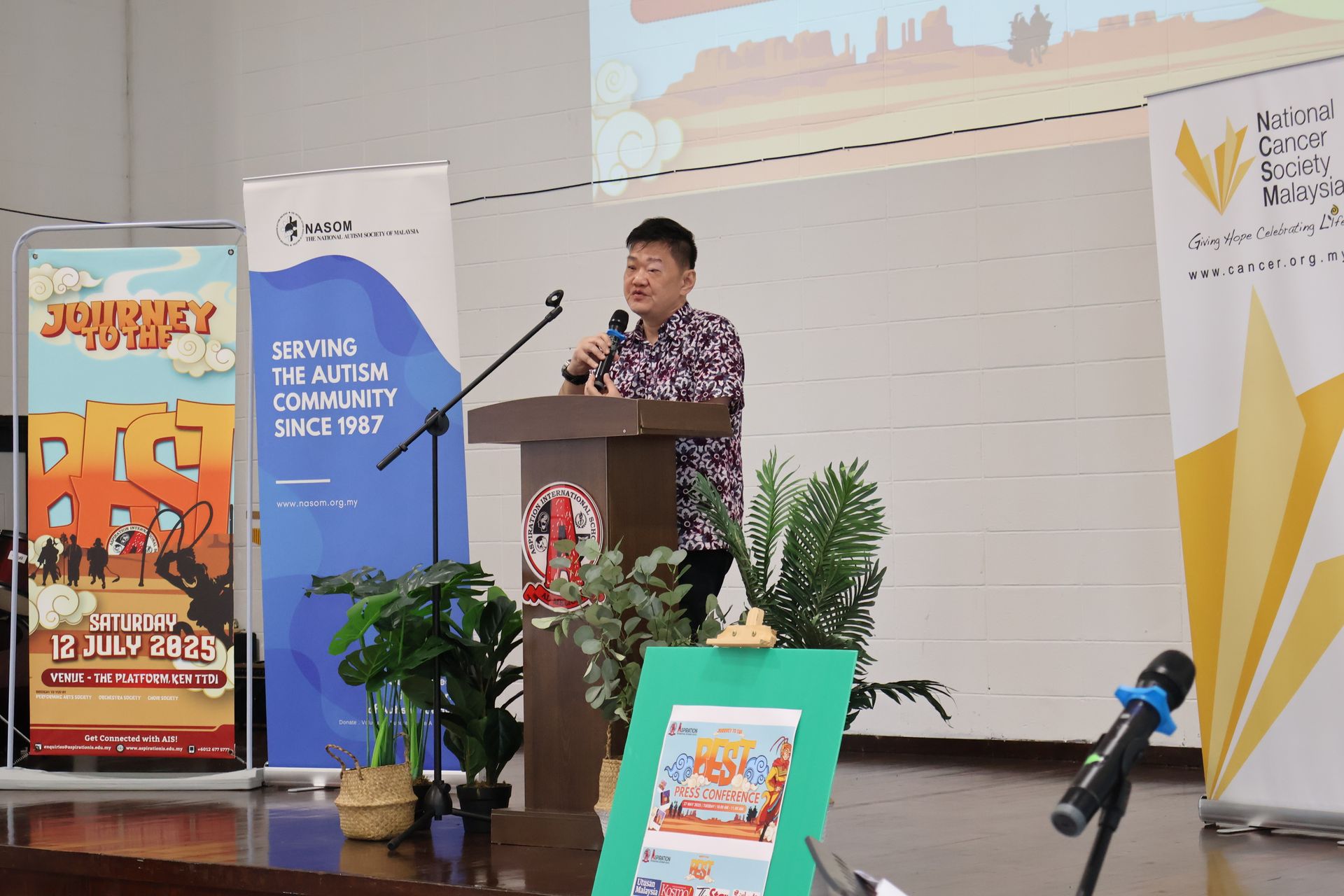 Man speaking at a podium, with banners and plants in a room.