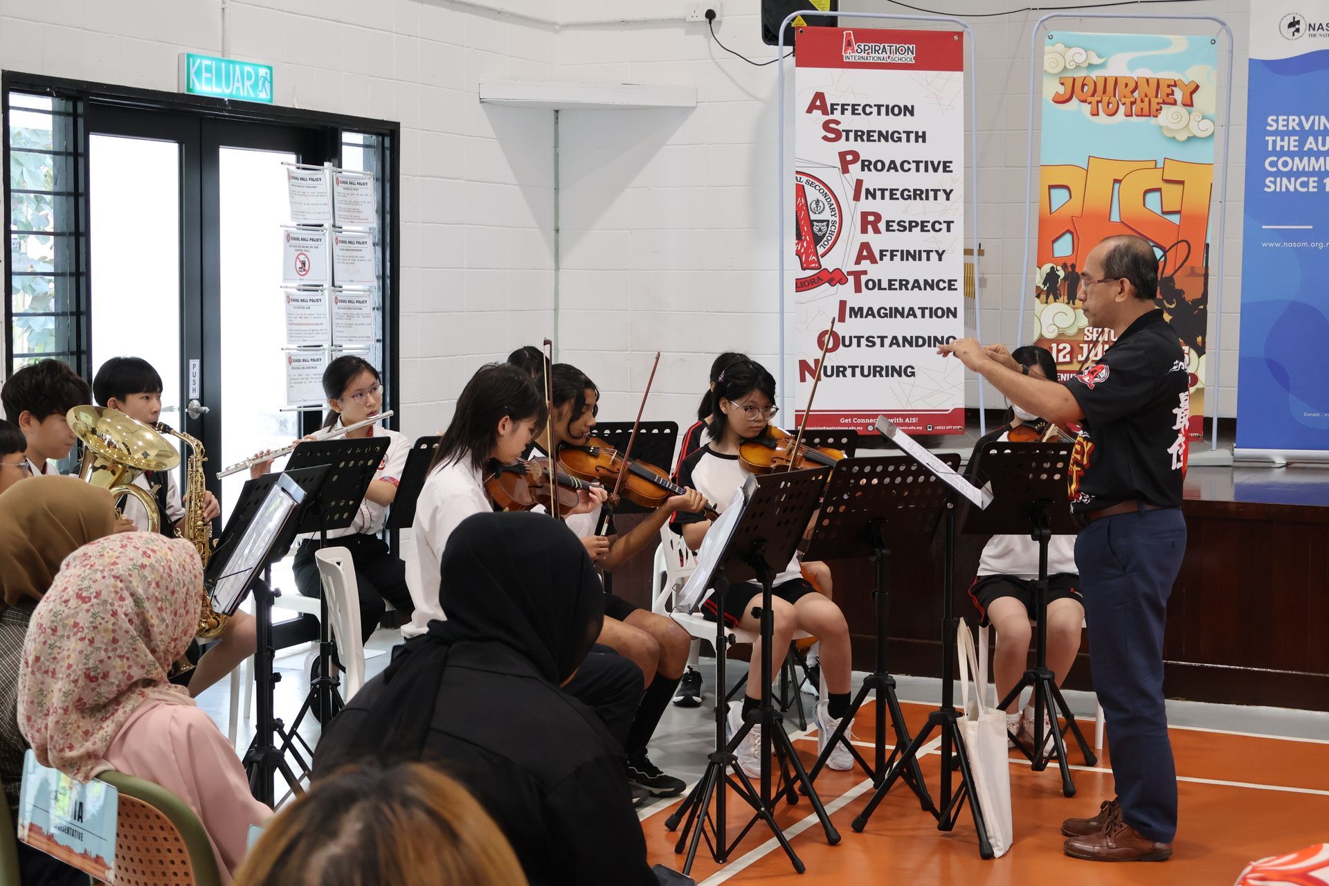 Orchestra performing in a hall; conductor directs string instruments. Audience watches.