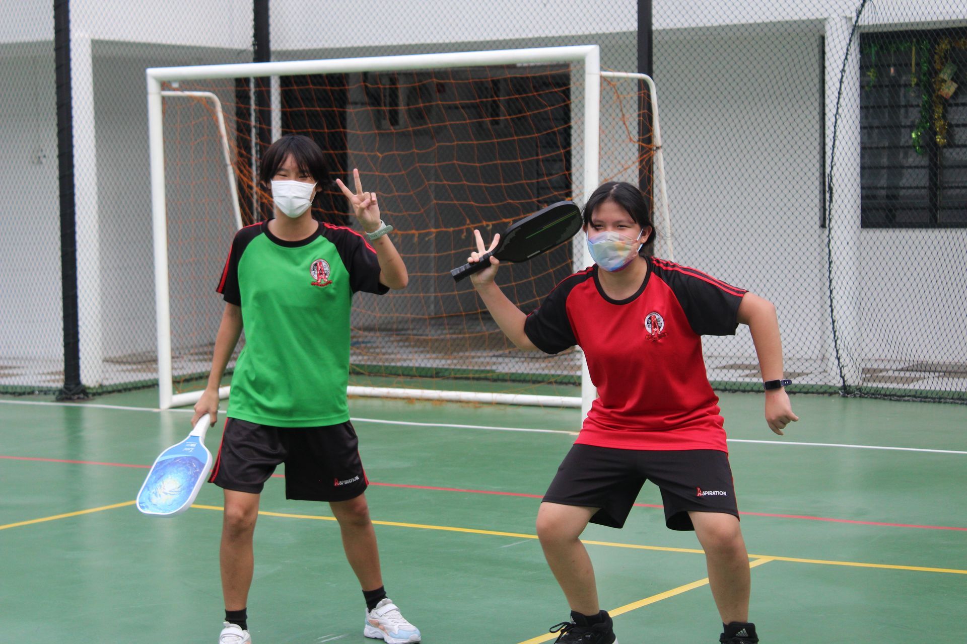 Two people with paddles in a sports court. One wears a green shirt, other a red shirt, both wearing masks.