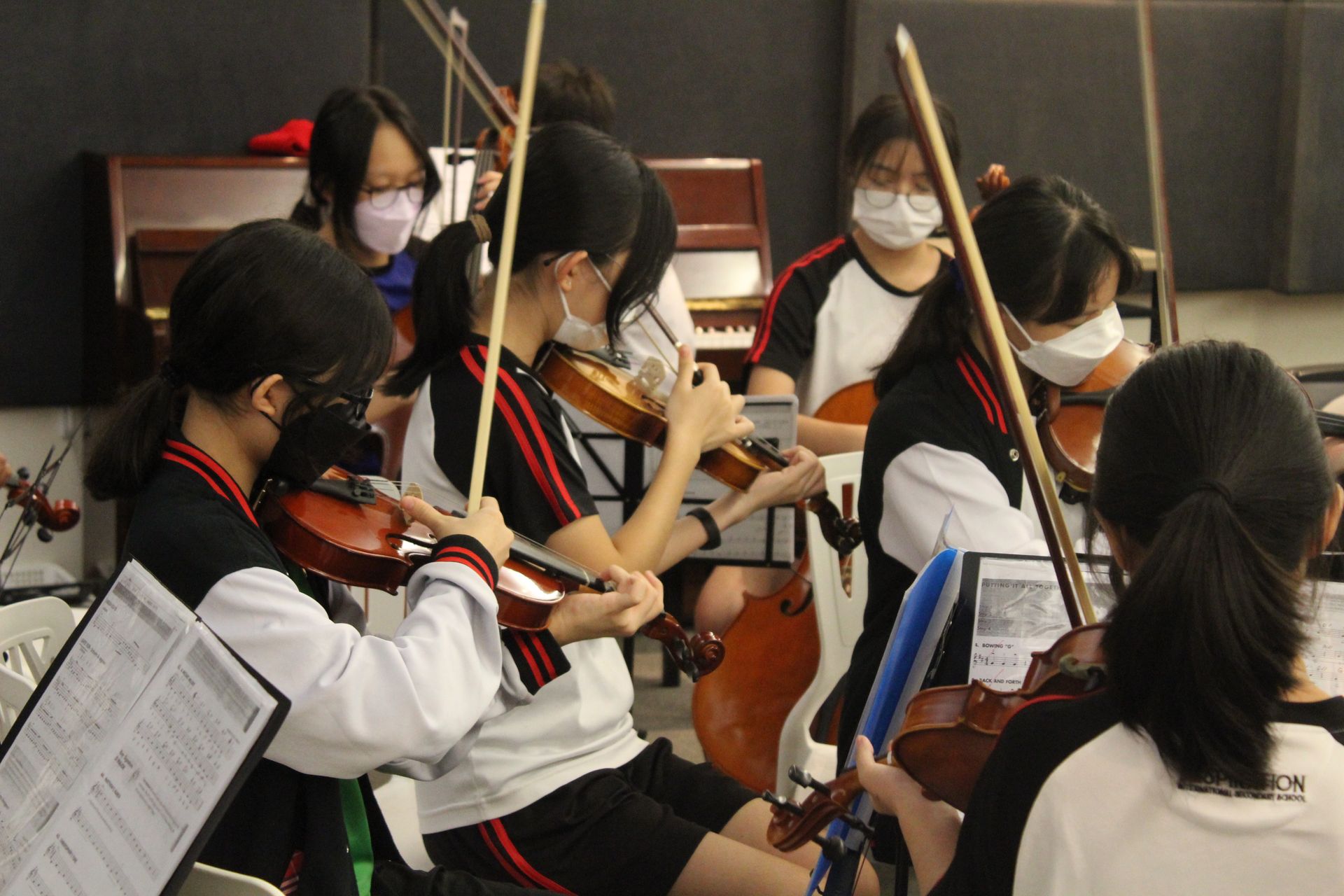 Students in masks playing violins in an orchestra, in a room with other instruments.