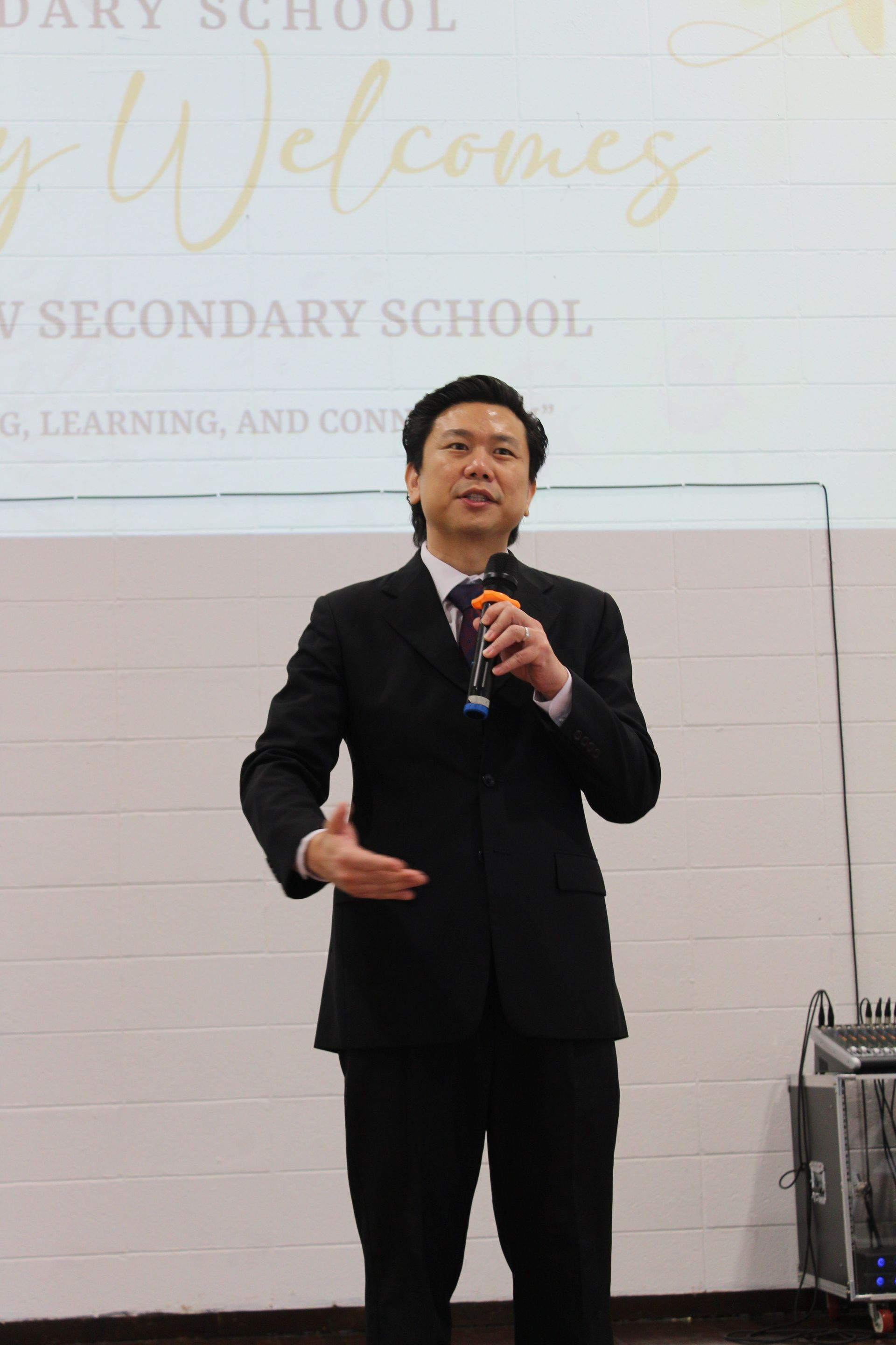 Man in a black suit speaks into a microphone, standing in front of a welcome banner.