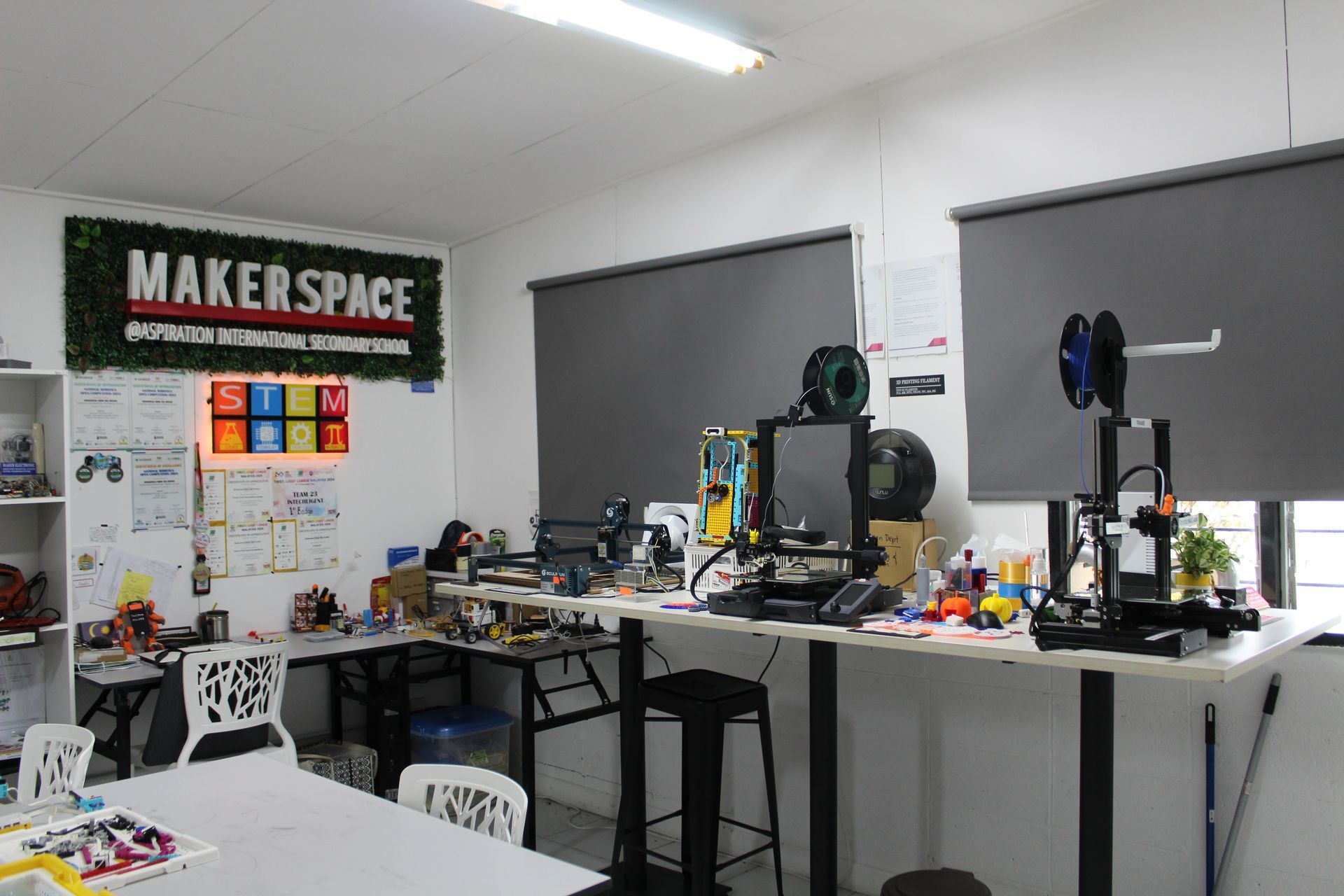 Maker space with 3D printers and work tables under grey window shades.