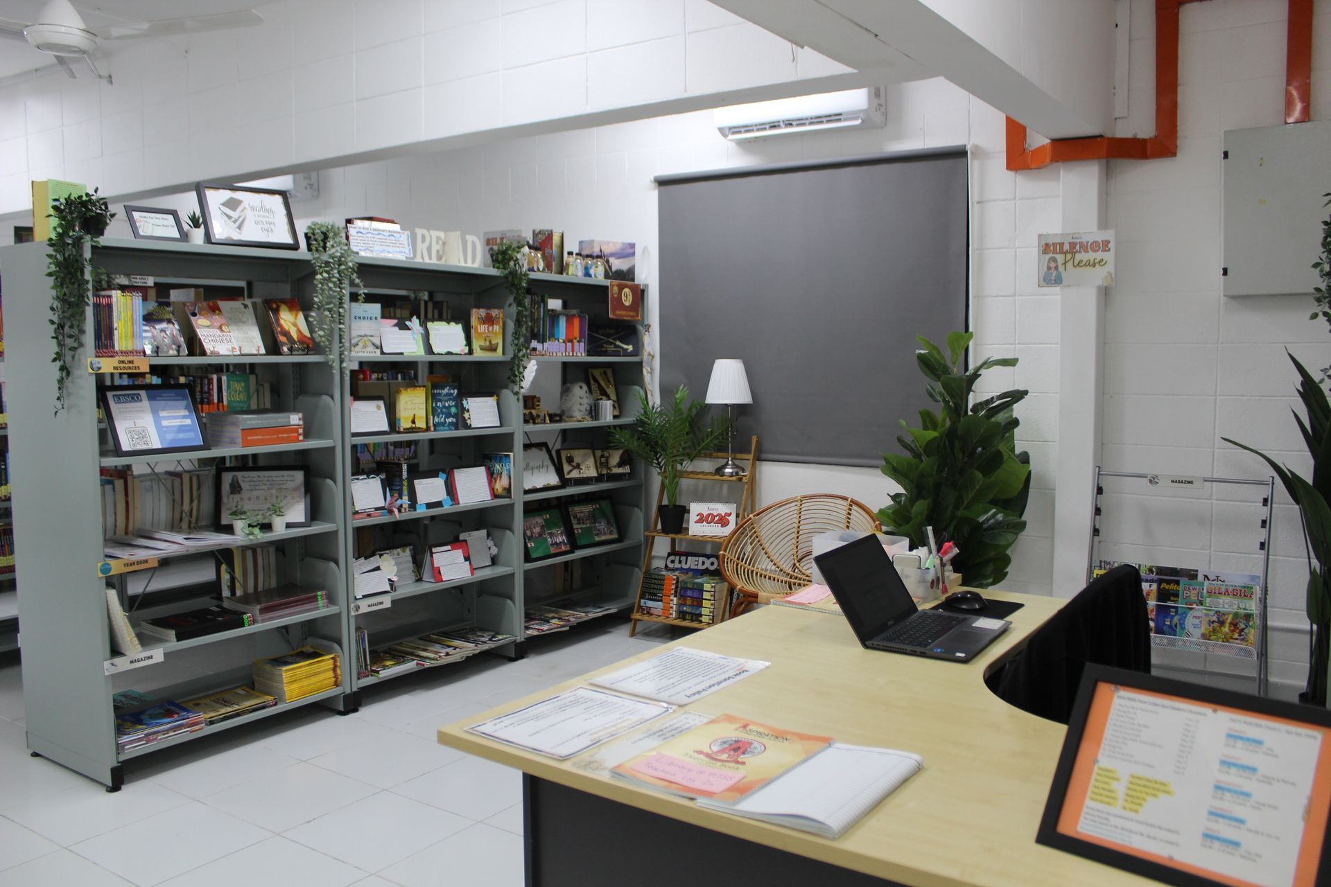 A library interior with bookshelves, a desk with a laptop, and decorative plants.