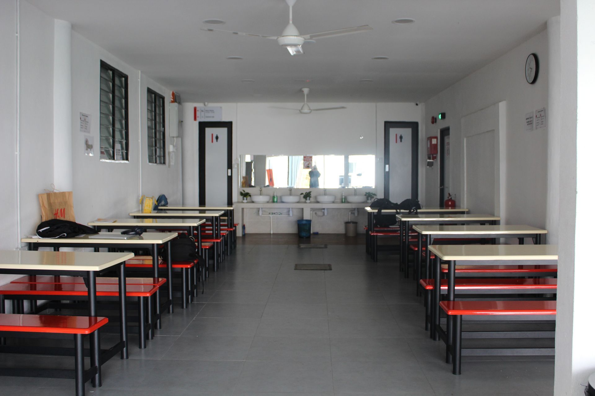 Dining hall with rows of tables and benches, white walls, and a washroom in the back.