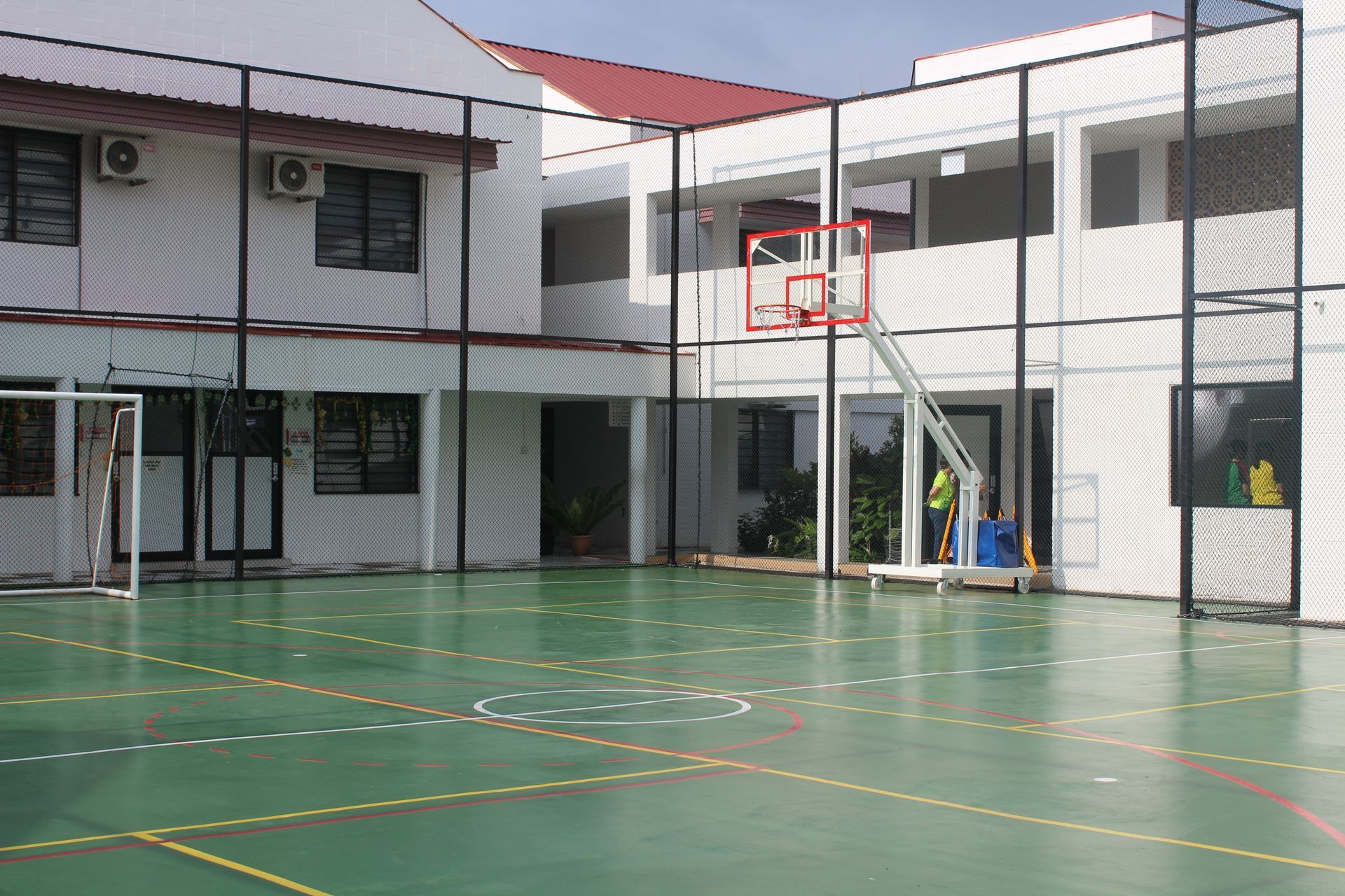 Outdoor basketball court with a hoop; building in background, green surface, fenced.