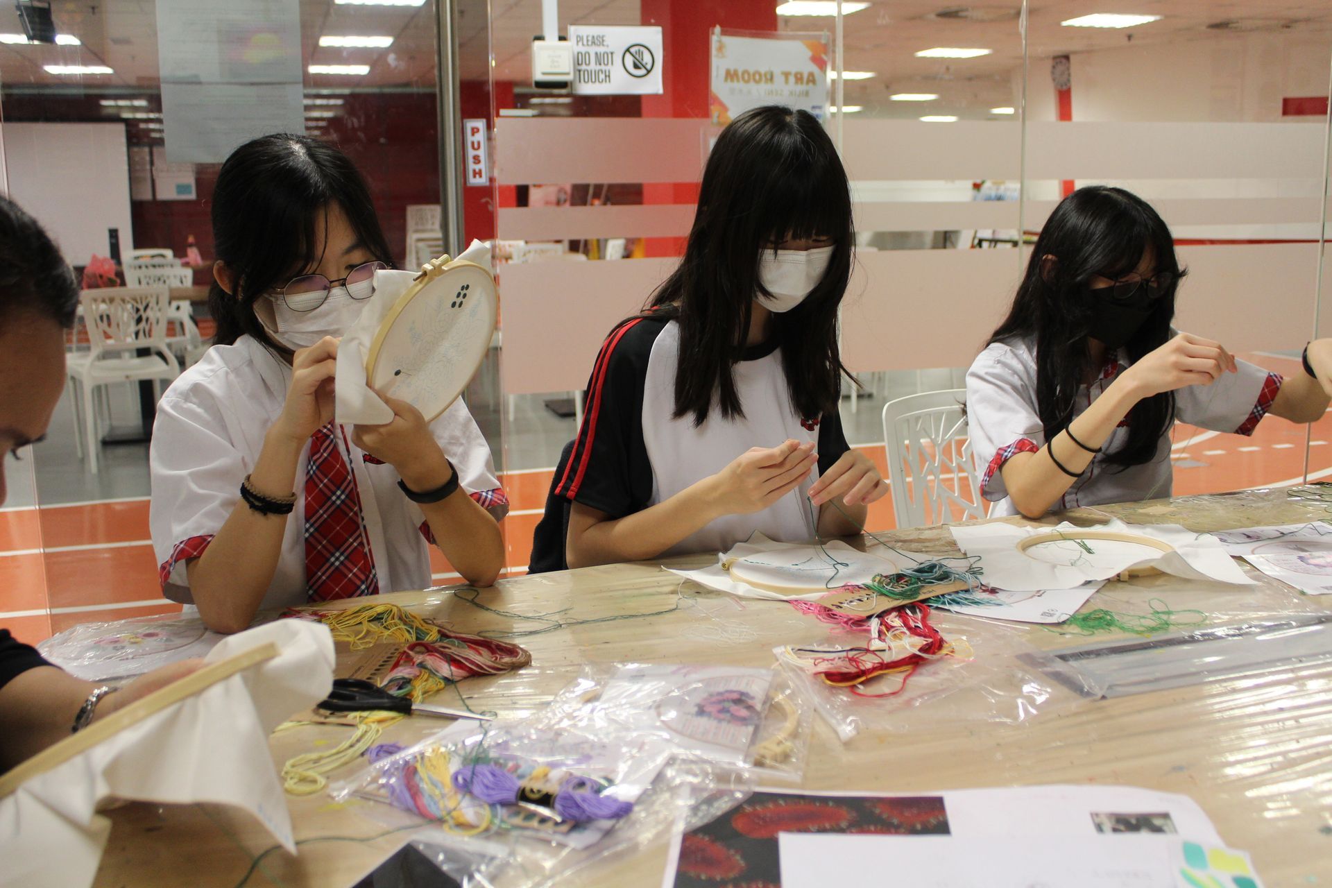 Students in masks embroidering at a table, inside a brightly lit room.