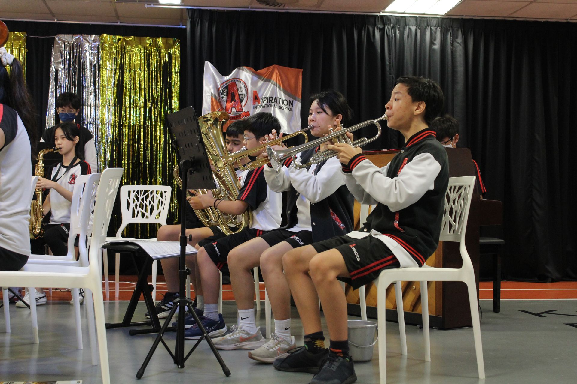 A band performs: trumpets, sax, tuba players seated on chairs in a room with a black backdrop.