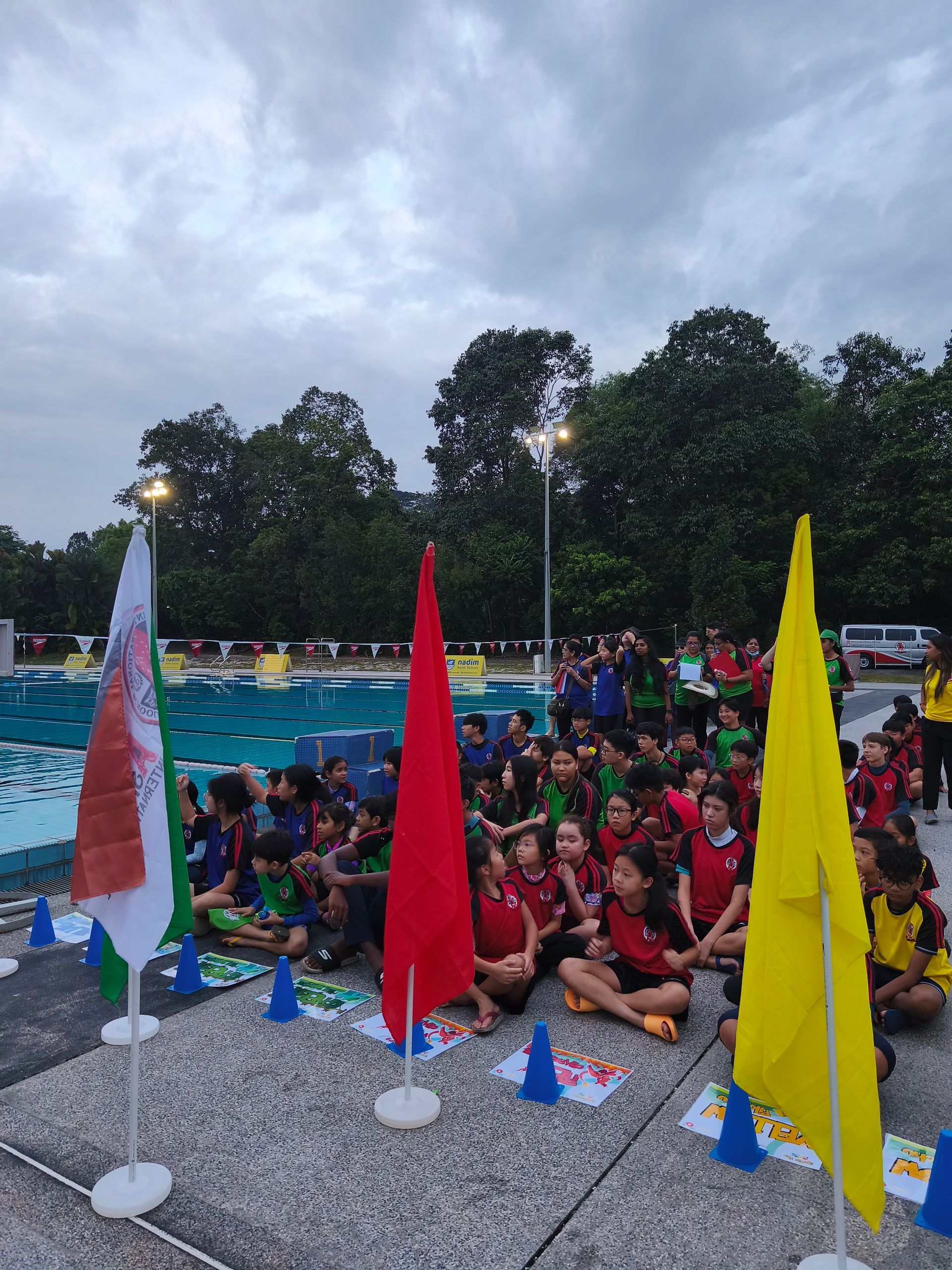 Children sitting on the ground near a swimming pool, flags of different colors in front of them.