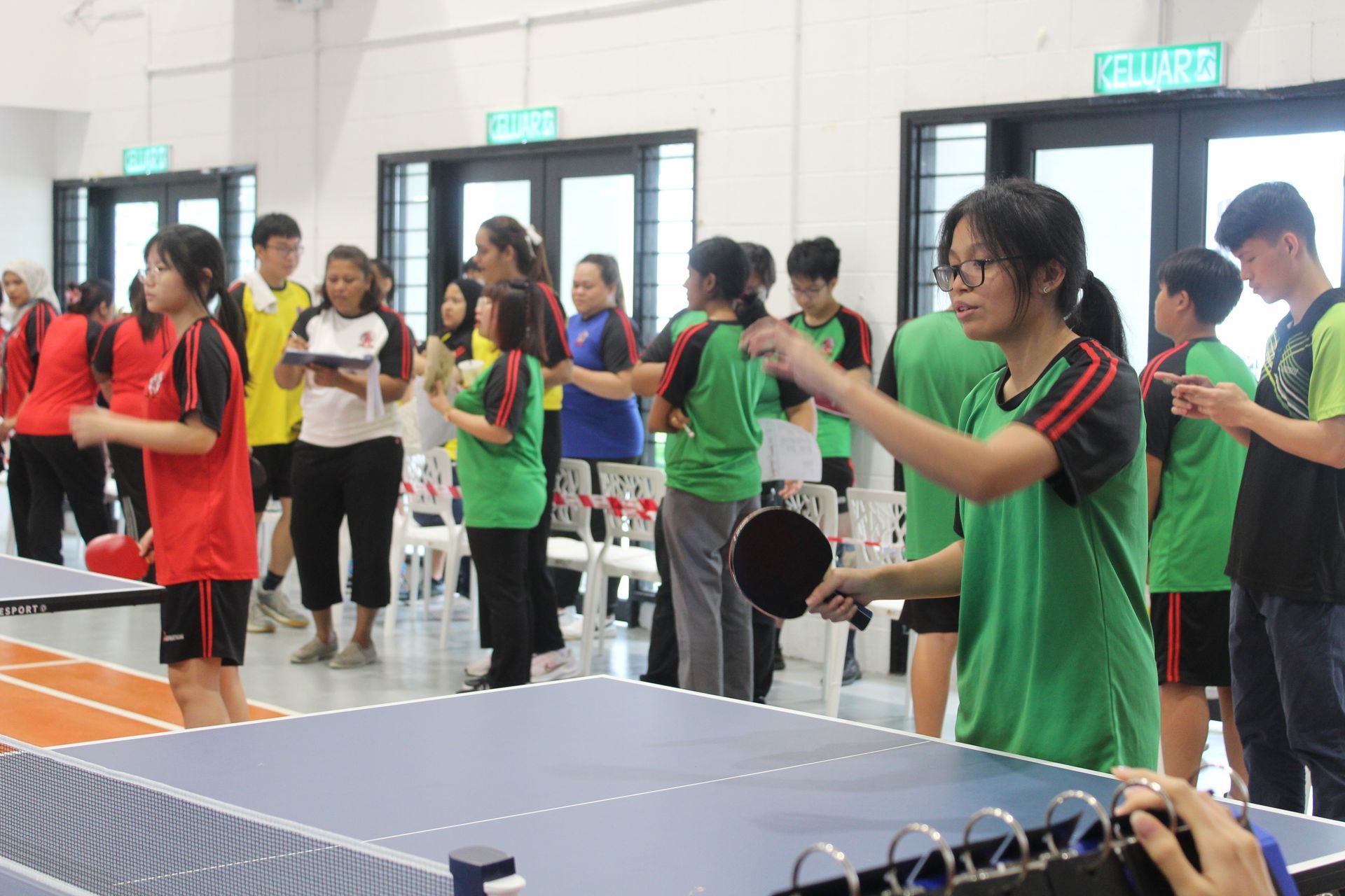 People playing table tennis in a brightly lit indoor sports hall. Players wear colored shirts, holding paddles. Others watch.
