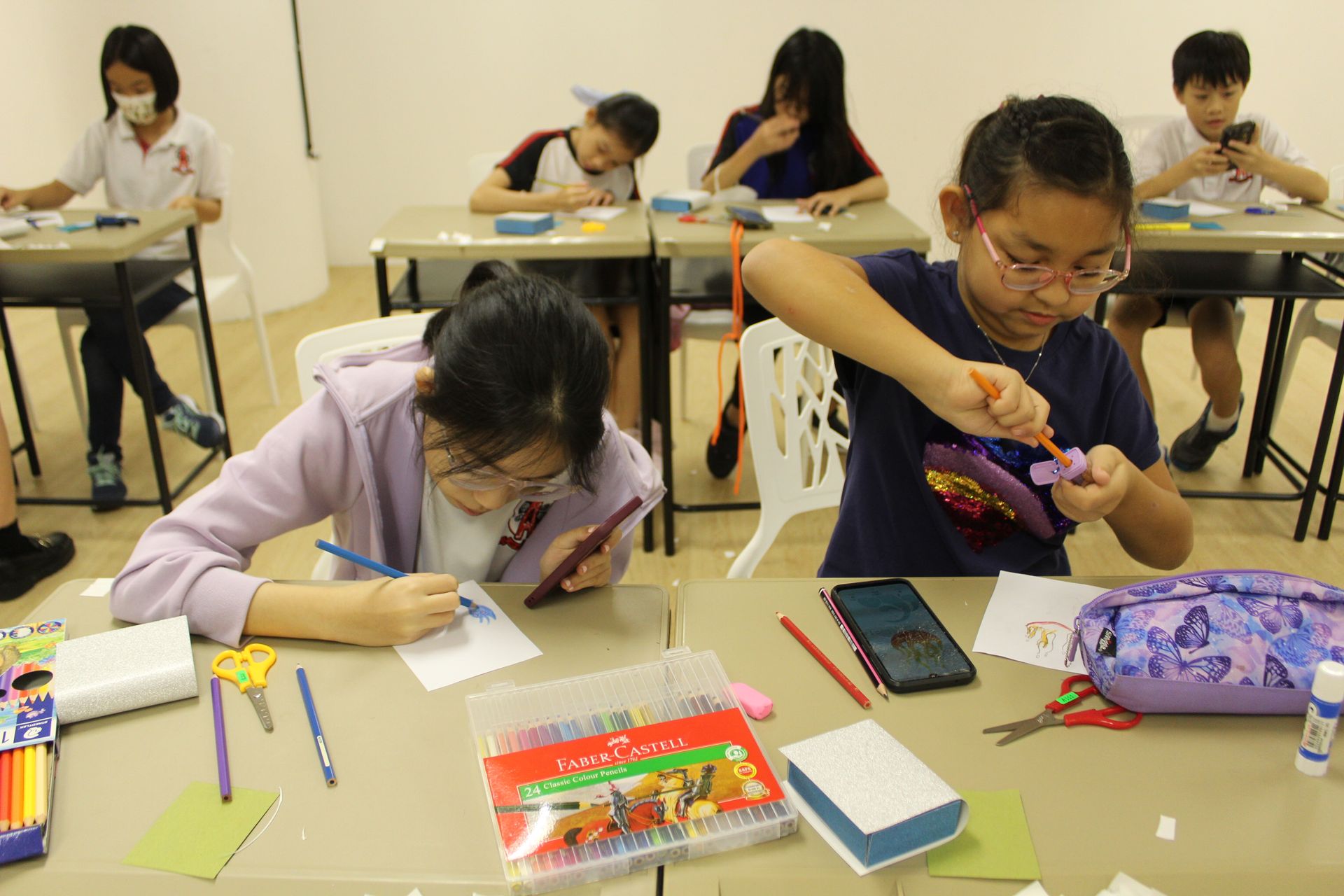 Children seated at desks, creating art. They are using pens, pencils, and scissors. One child uses a glue stick.