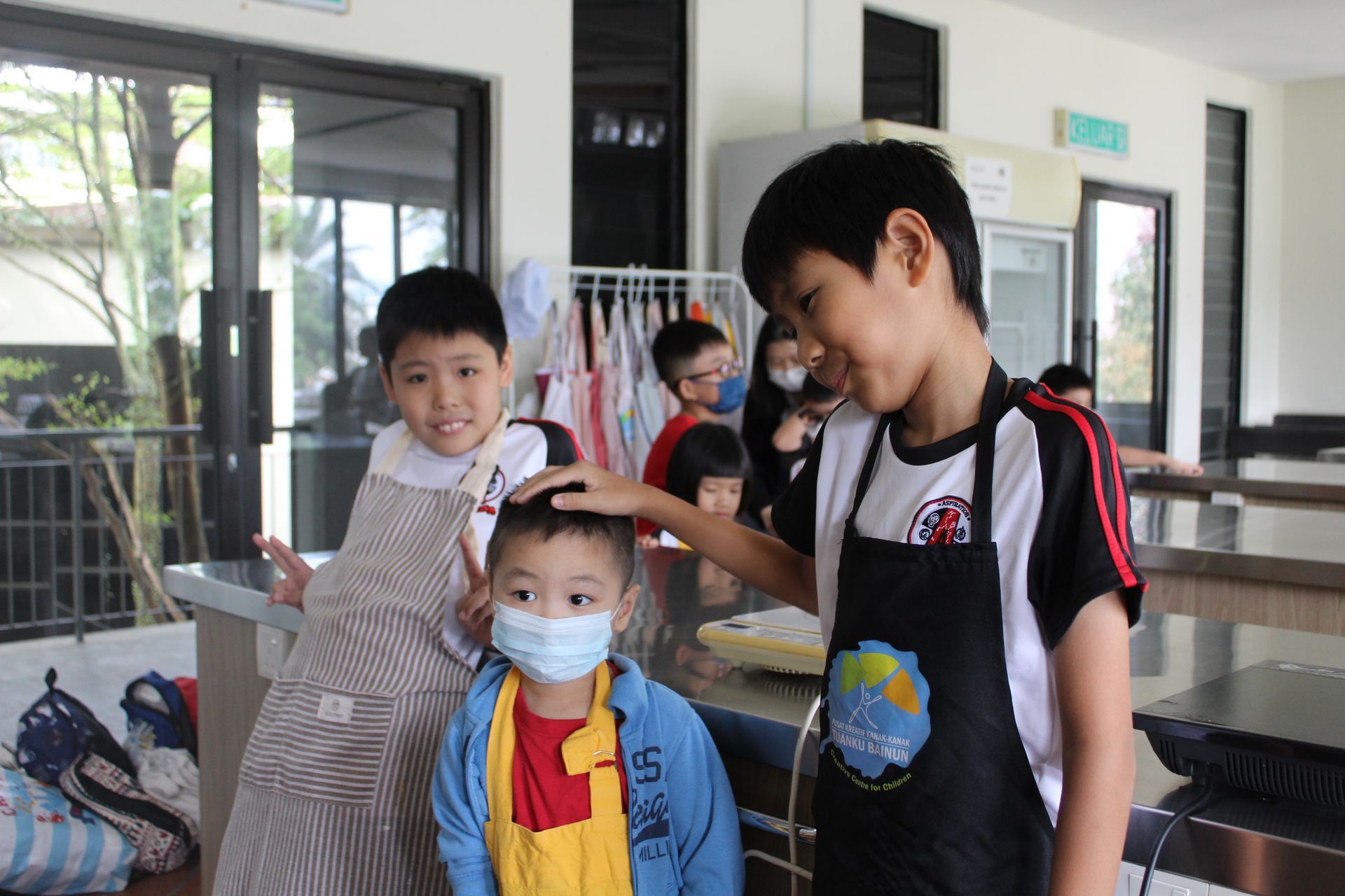 Children in aprons in a kitchen, one is touching another's head, smiling.