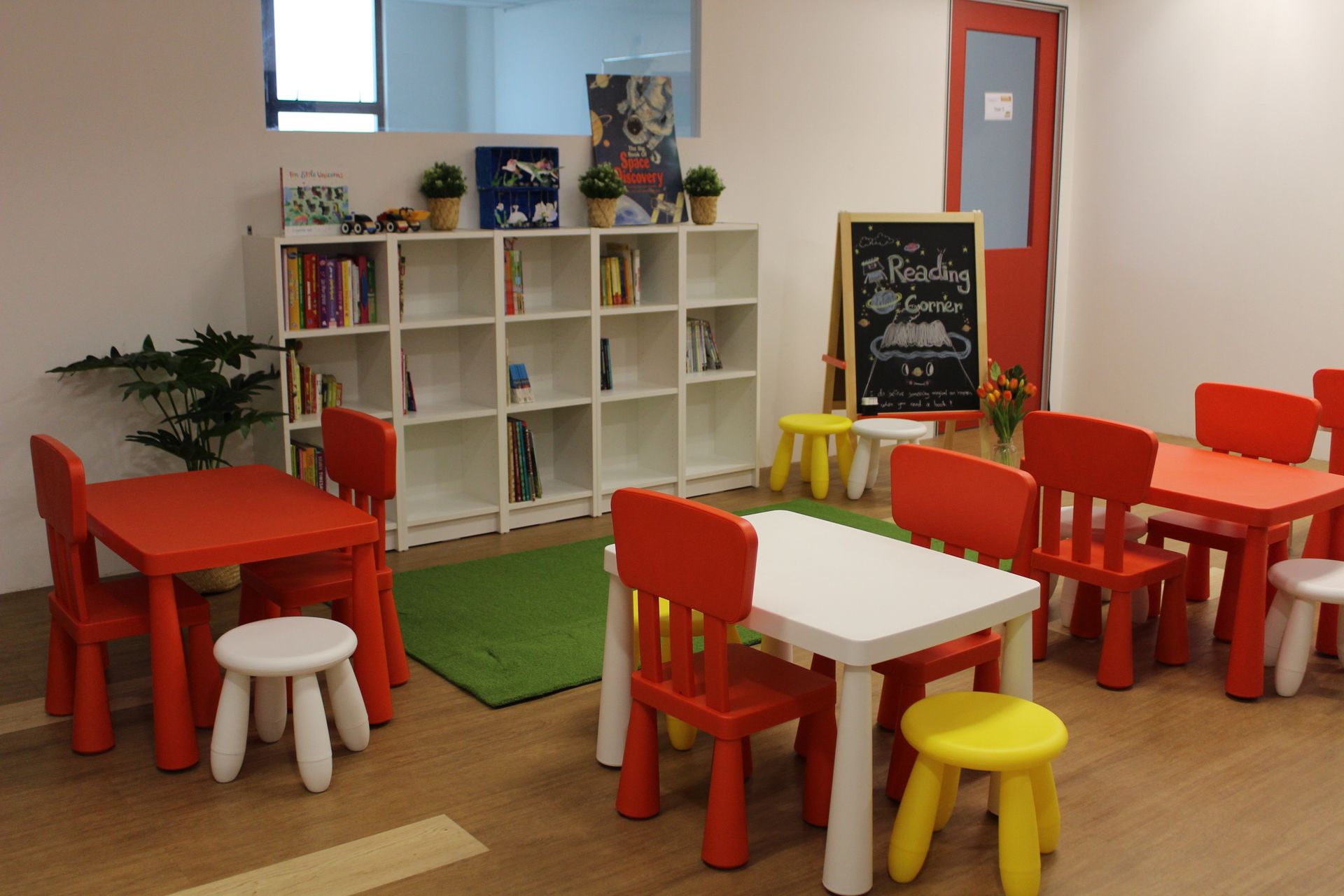 A brightly lit children's play area with red and white tables, colorful stools, bookshelves, and a chalkboard.