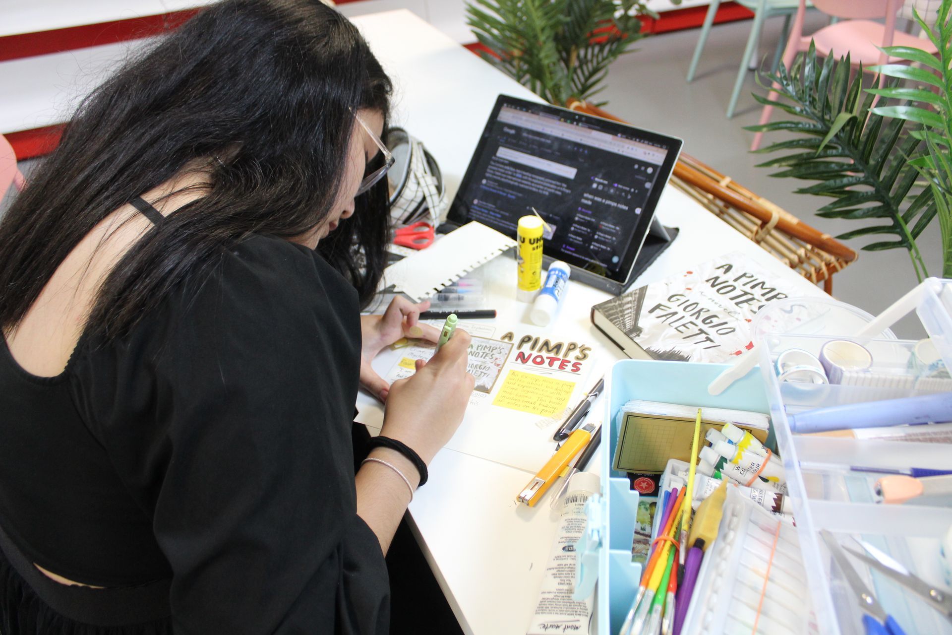 Two people working at a desk with laptop, art supplies, and a handwritten sign.