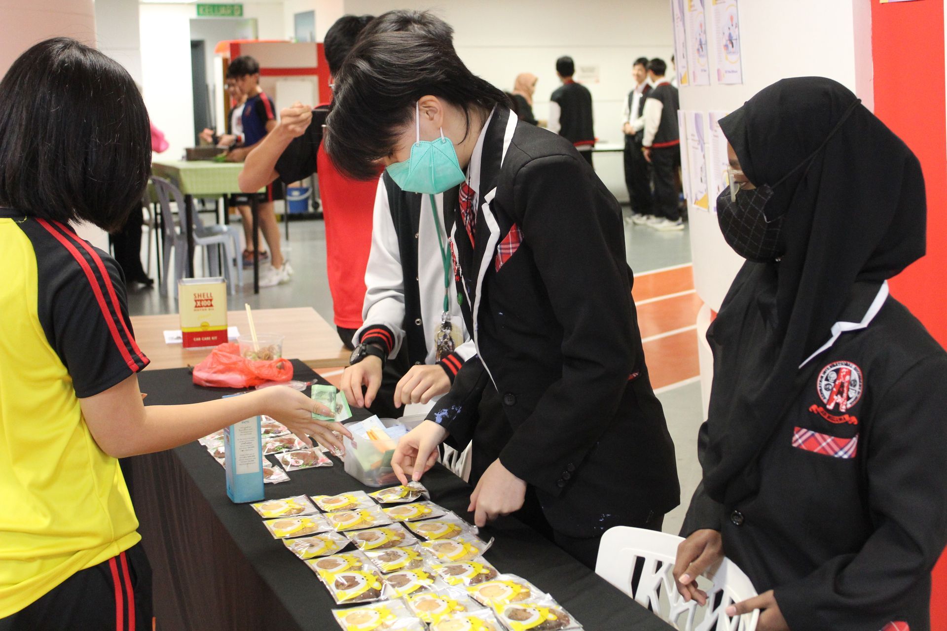 Students at a table, selling items; one wearing a mask, another is handing out something. Indoors, brightly lit.