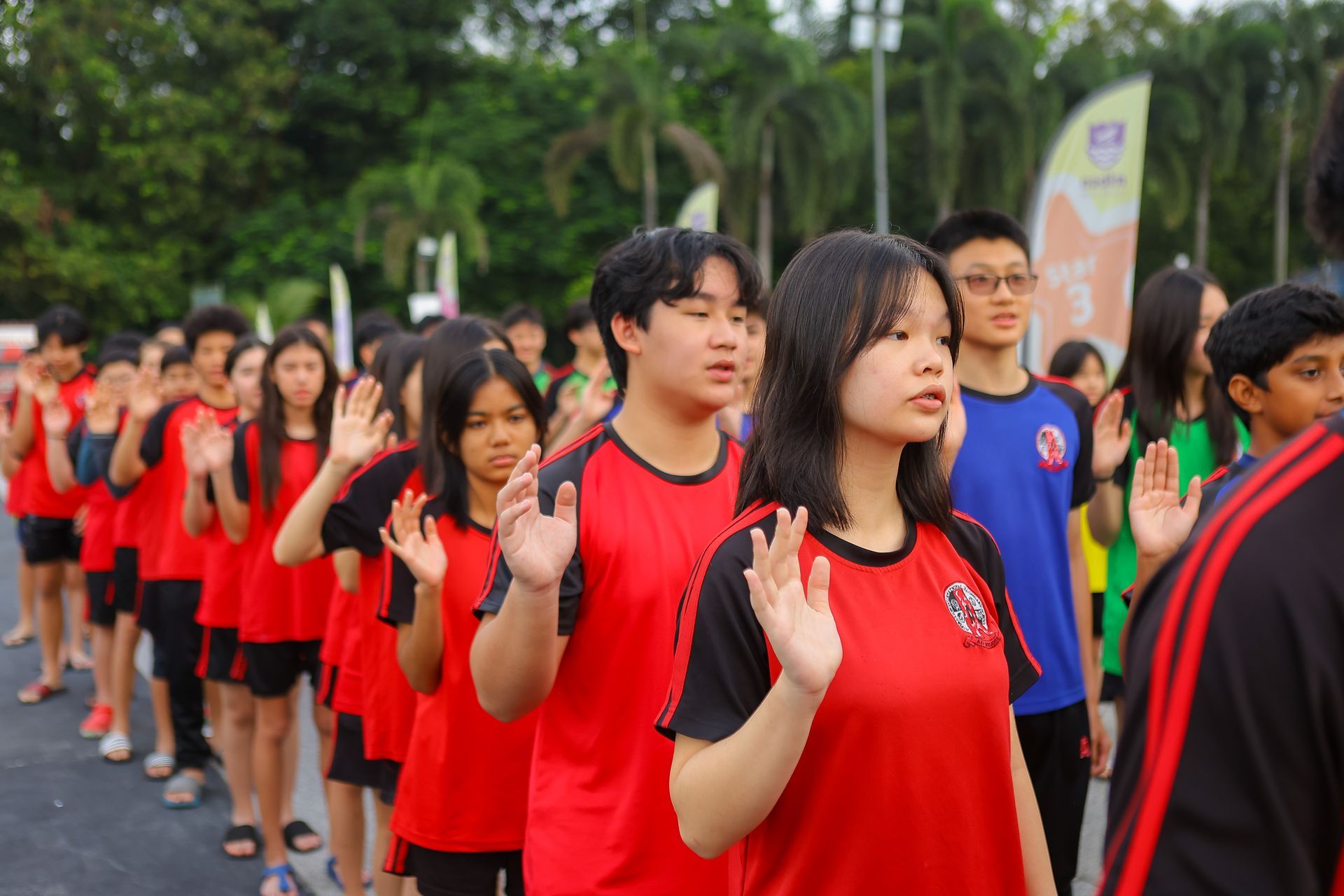 Students in red shirts raise their hands outdoors.