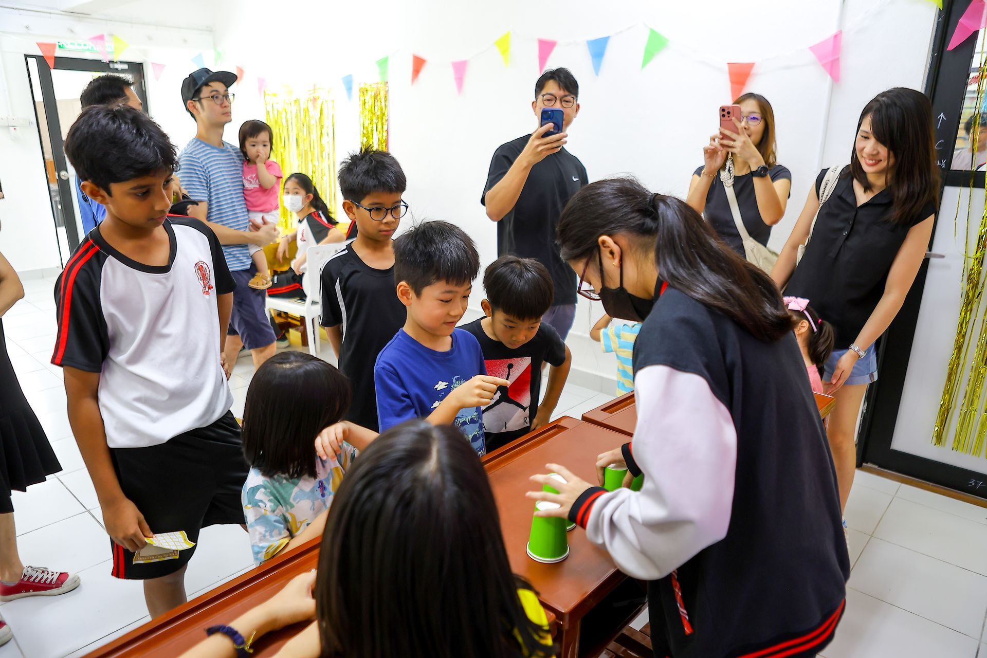 People playing a game with cups on a table, others watching. Flags and decorations are in a room.