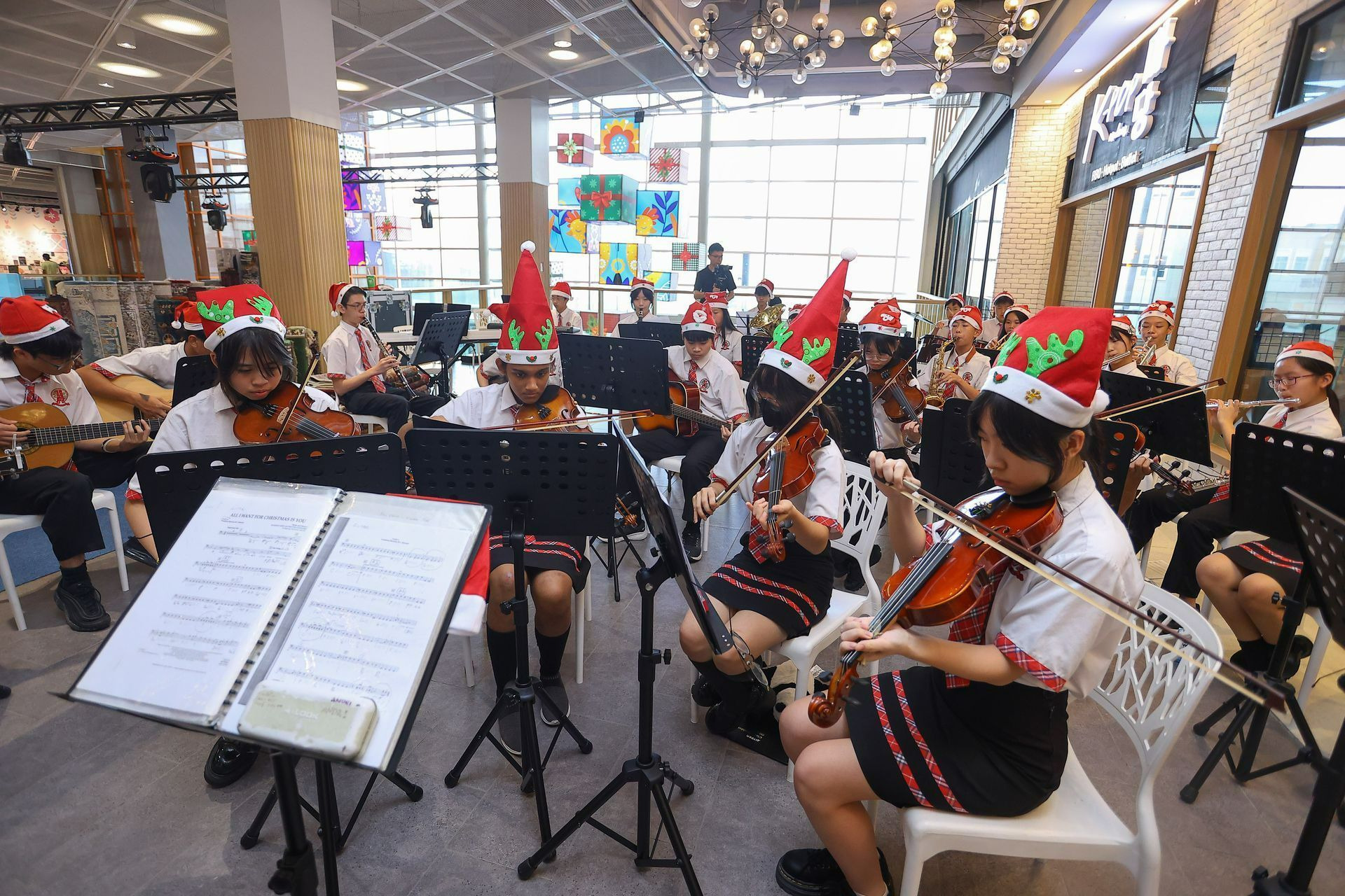 An orchestra in Santa hats plays violins in a brightly lit indoor space with Christmas decorations.