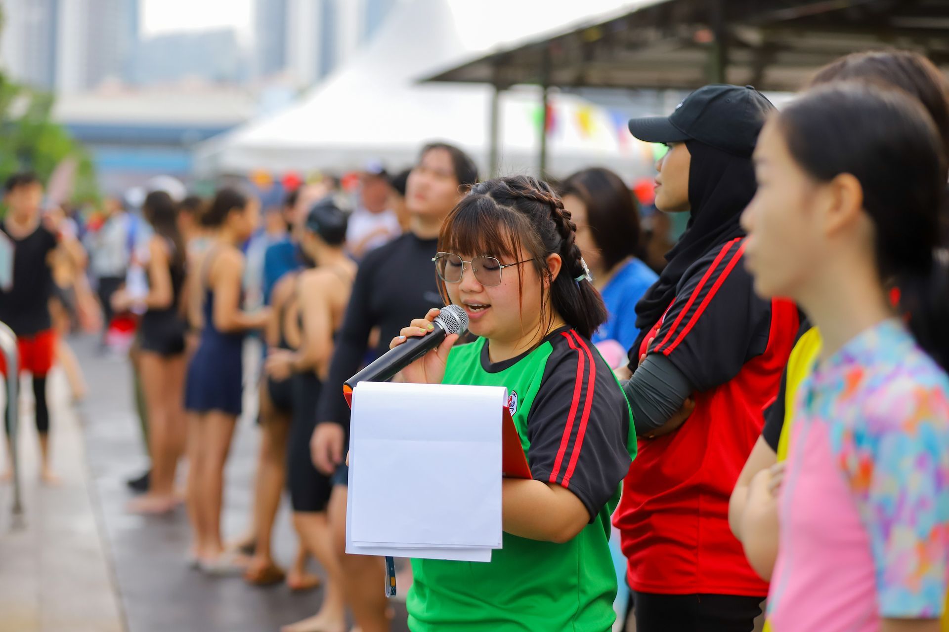 A person with Down syndrome speaks into a microphone, holding paper, at an outdoor event near a pool.