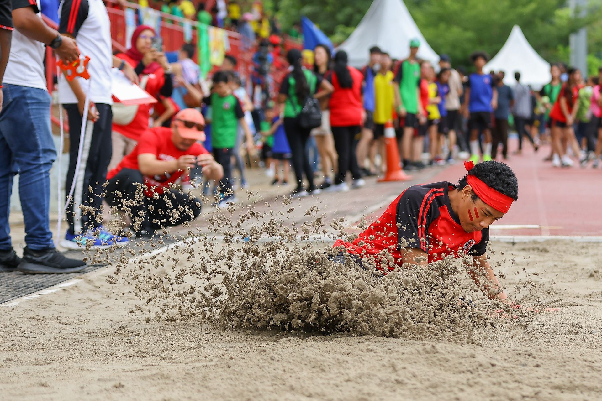 Athlete lands in sandpit during a long jump competition, surrounded by spectators.