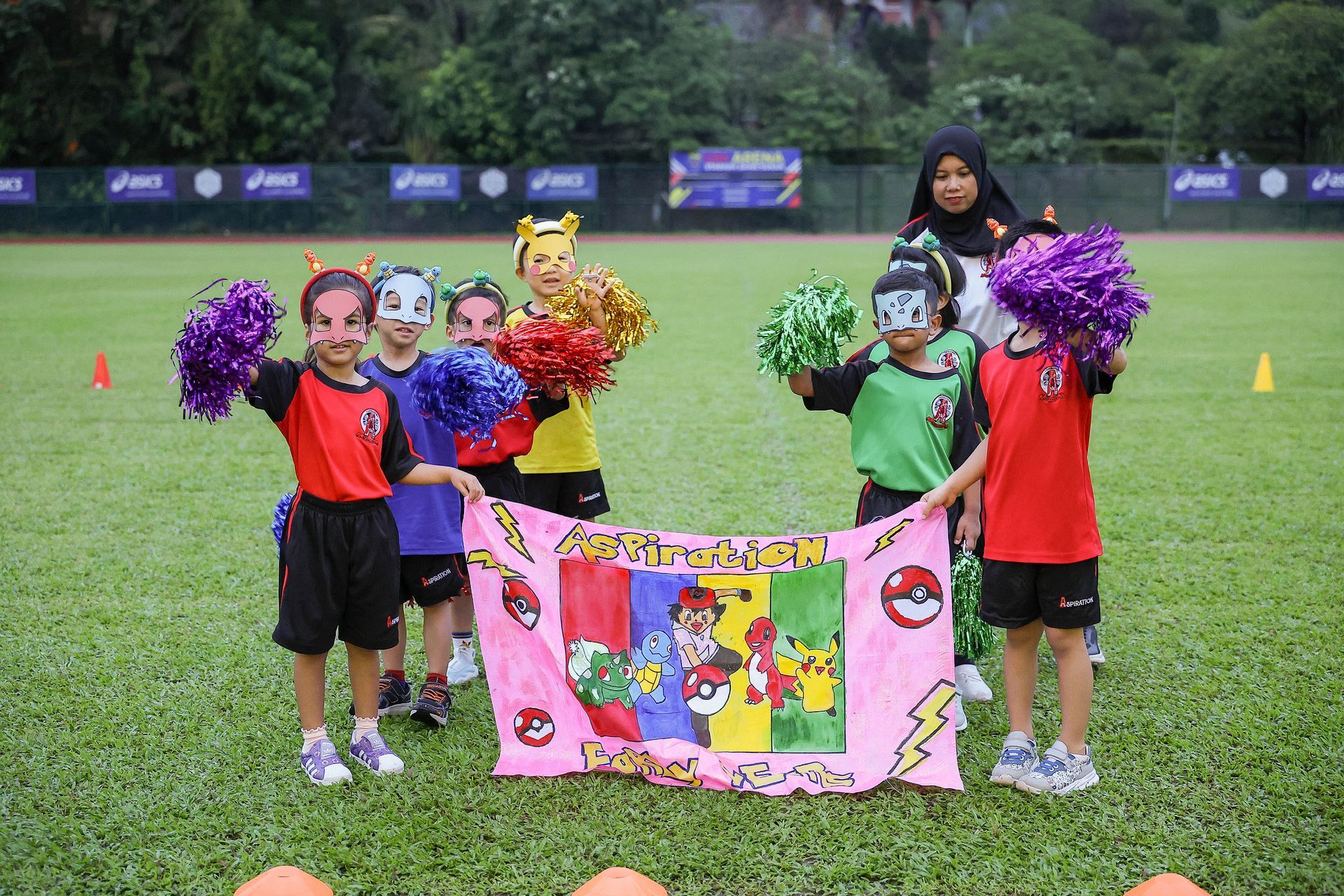 Children in costumes with pom-poms hold a banner on a grass field; an adult in a hijab watches.