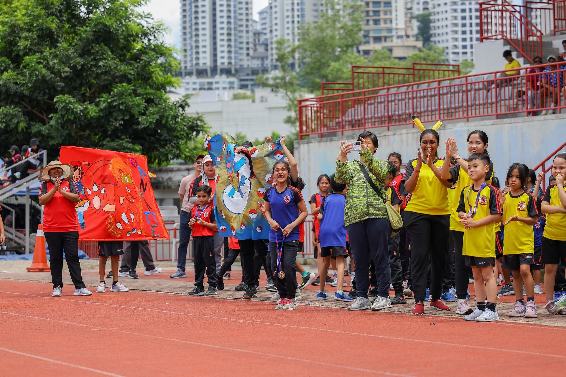 Children in colorful outfits parade at a track, holding decorations. Buildings are in the background.