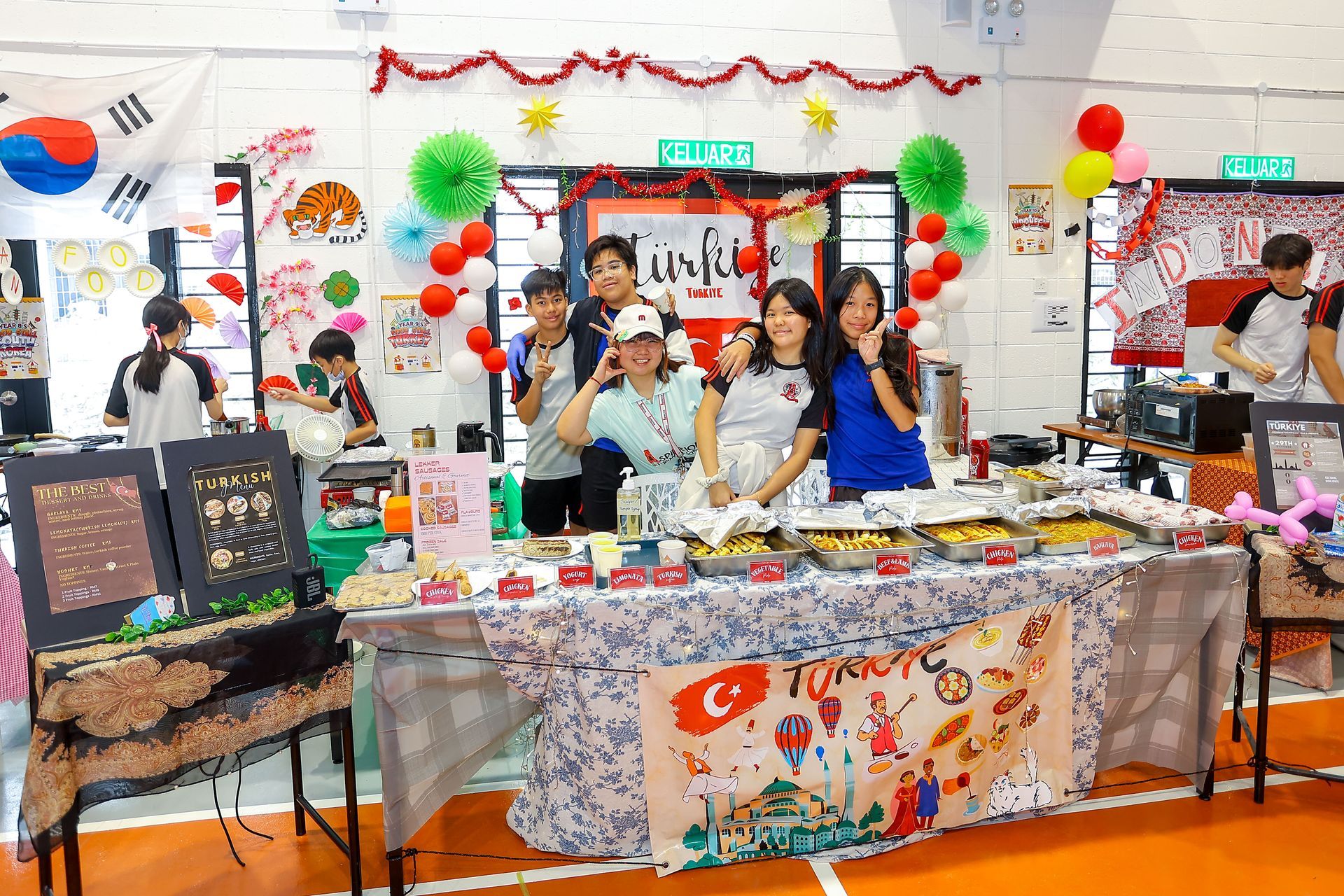 School festival booth. Group of teens smiling behind table with Turkish flag and food, decorations overhead.