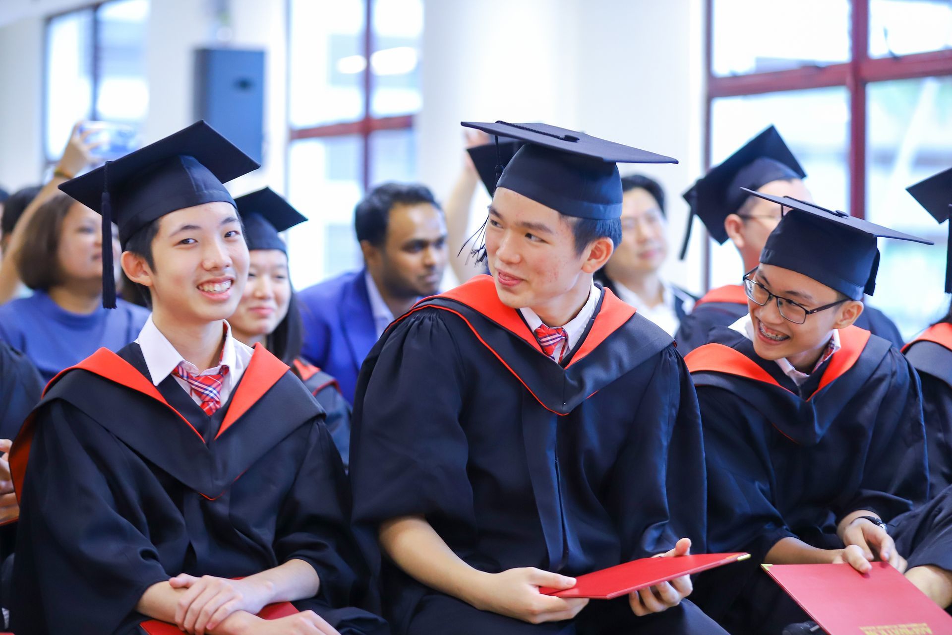 Graduates in black gowns and mortarboards, holding red folders, smiling at a ceremony.