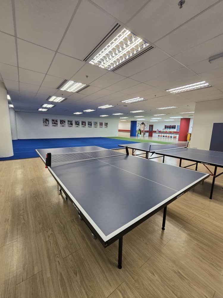 Ping pong tables in a brightly lit room with a blue floor and other tables visible in the background.