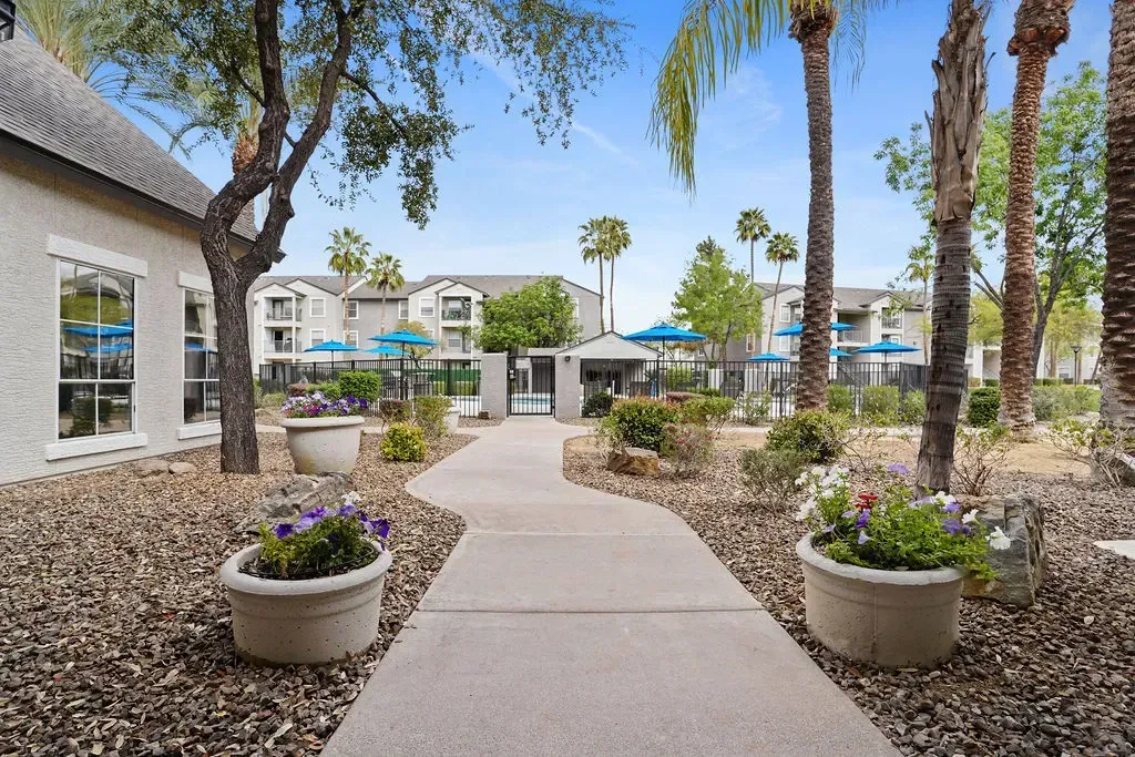 Courtyard of an apartment complex with a paved path, palm trees, planters, and a fenced pool.