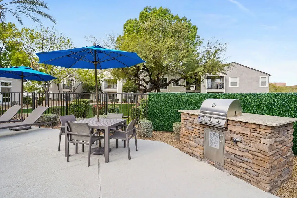 Outdoor communal grilling area with table, chairs, and blue umbrellas.