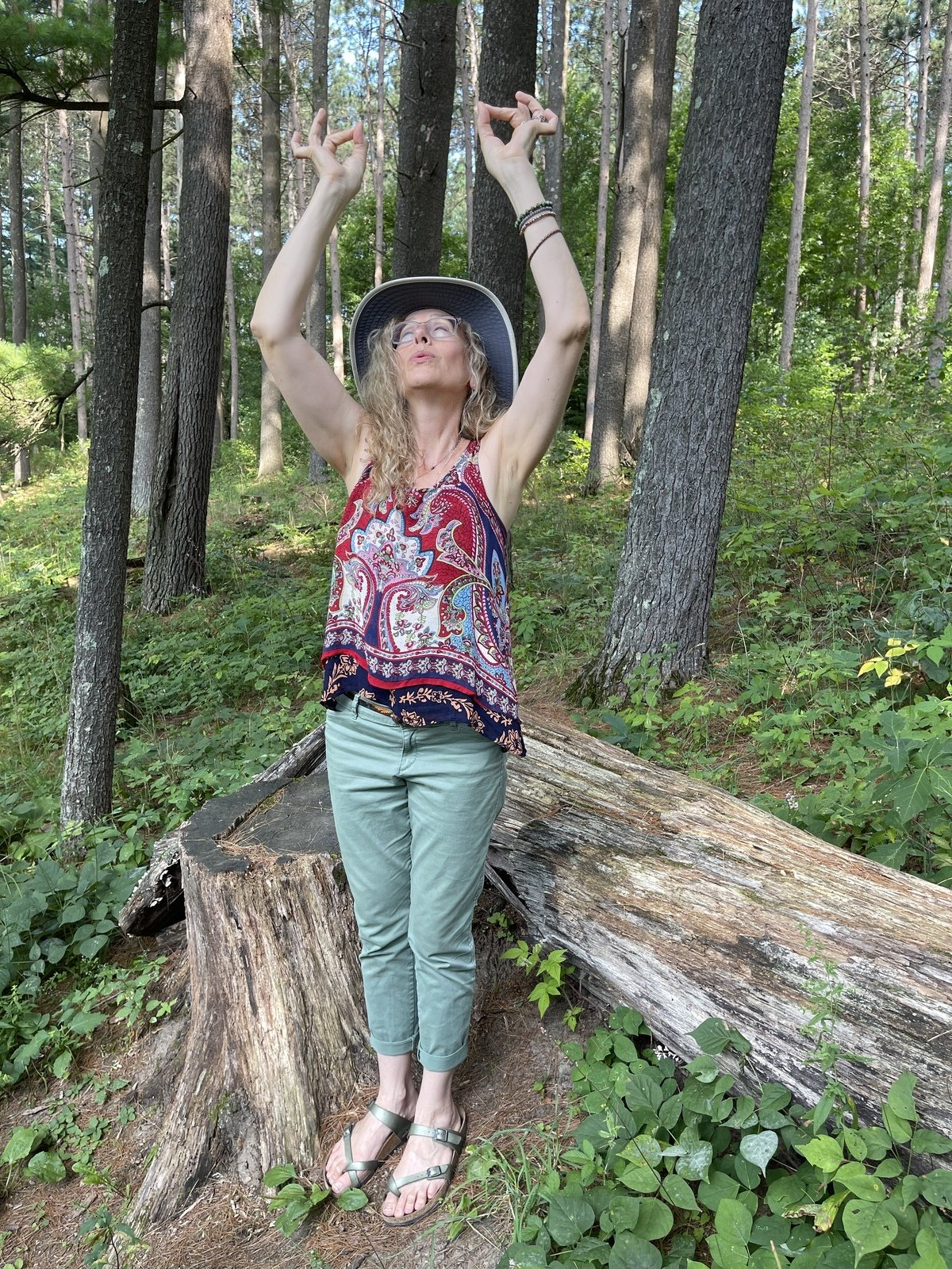 A woman is standing on a tree stump in the woods with her arms in the air.
