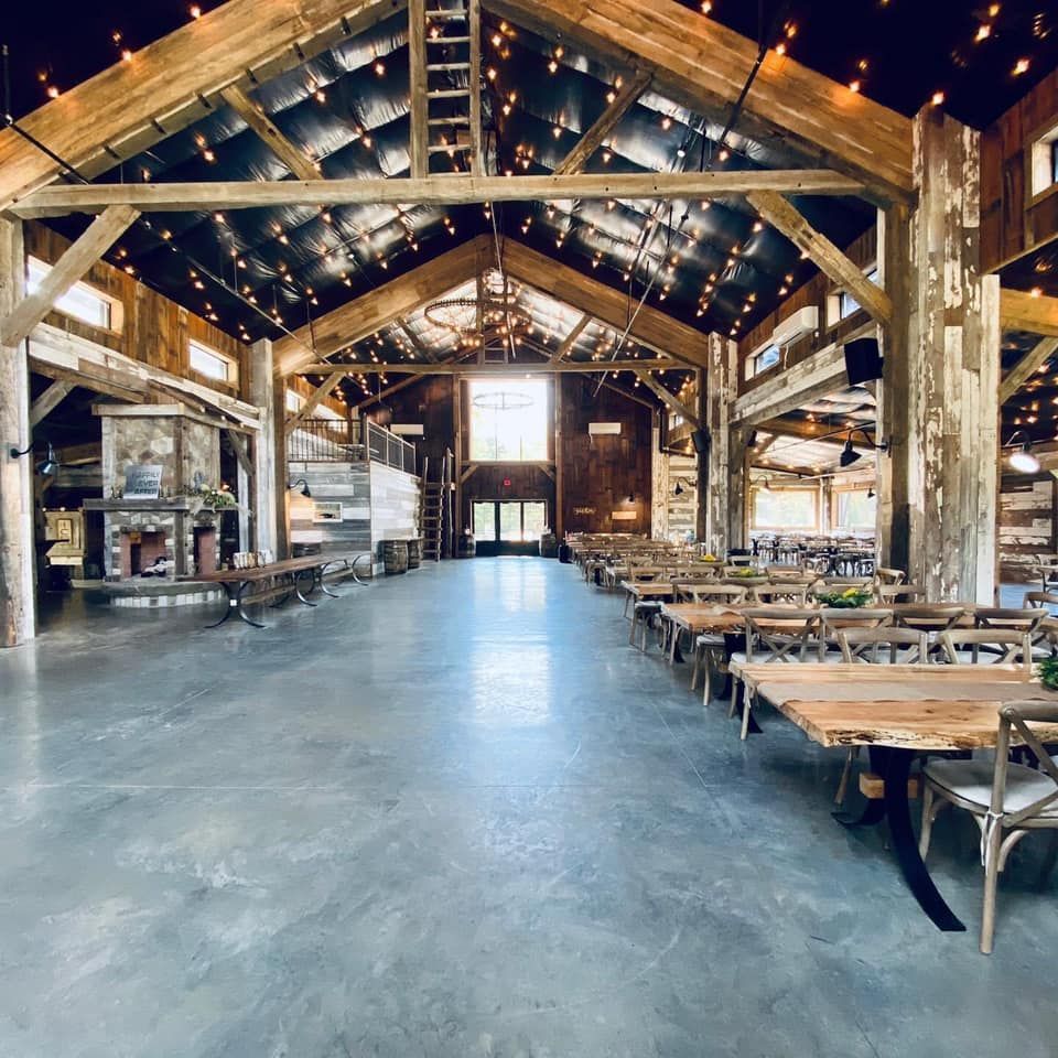 Spacious barn interior with wooden beams, concrete floor, tables, chairs, and string lights.