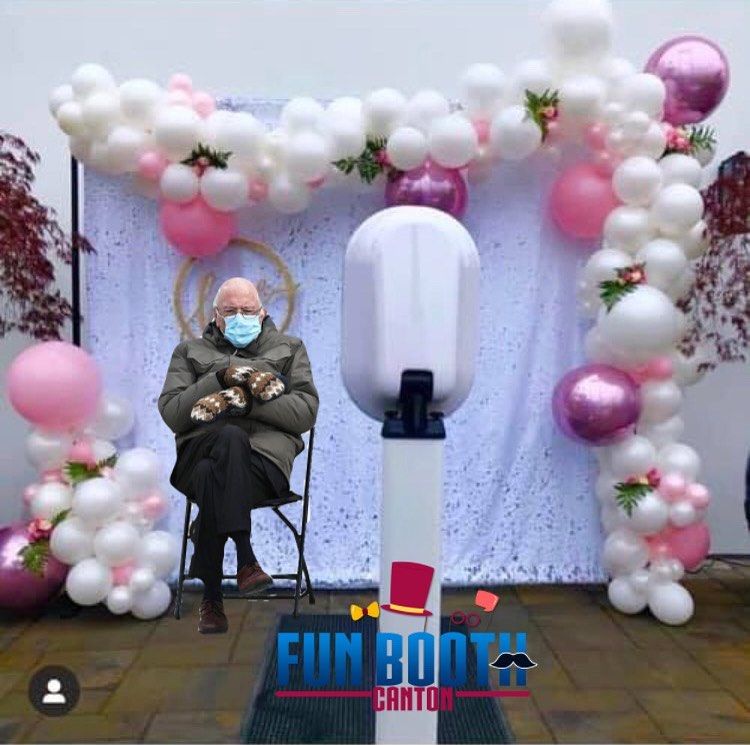 Bernie Sanders sits in chair, wearing mittens and a mask, next to a photo booth decorated with pink and white balloons.