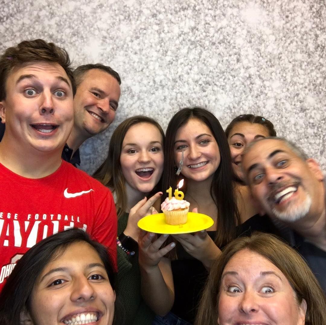 Group of eight people smiling, celebrating a birthday, holding a cupcake with a lit candle, against a textured wall.