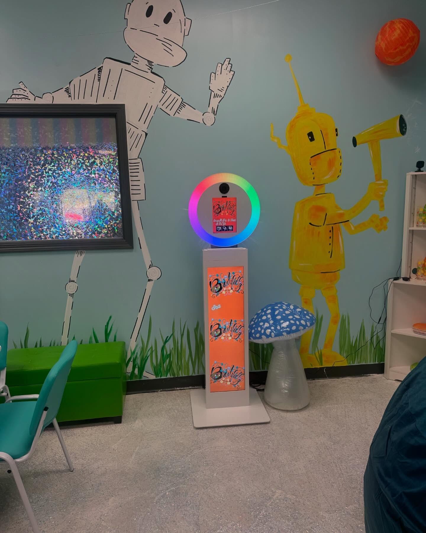 Photo booth with a rainbow-lit ring, in front of a colorful mural featuring cartoon figures and a mushroom.