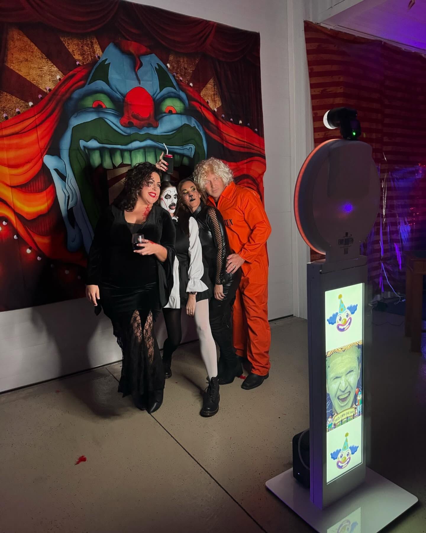 People pose for a photo near a clown backdrop. A photo booth sits to the side. They wear costumes and have expressions of fun.