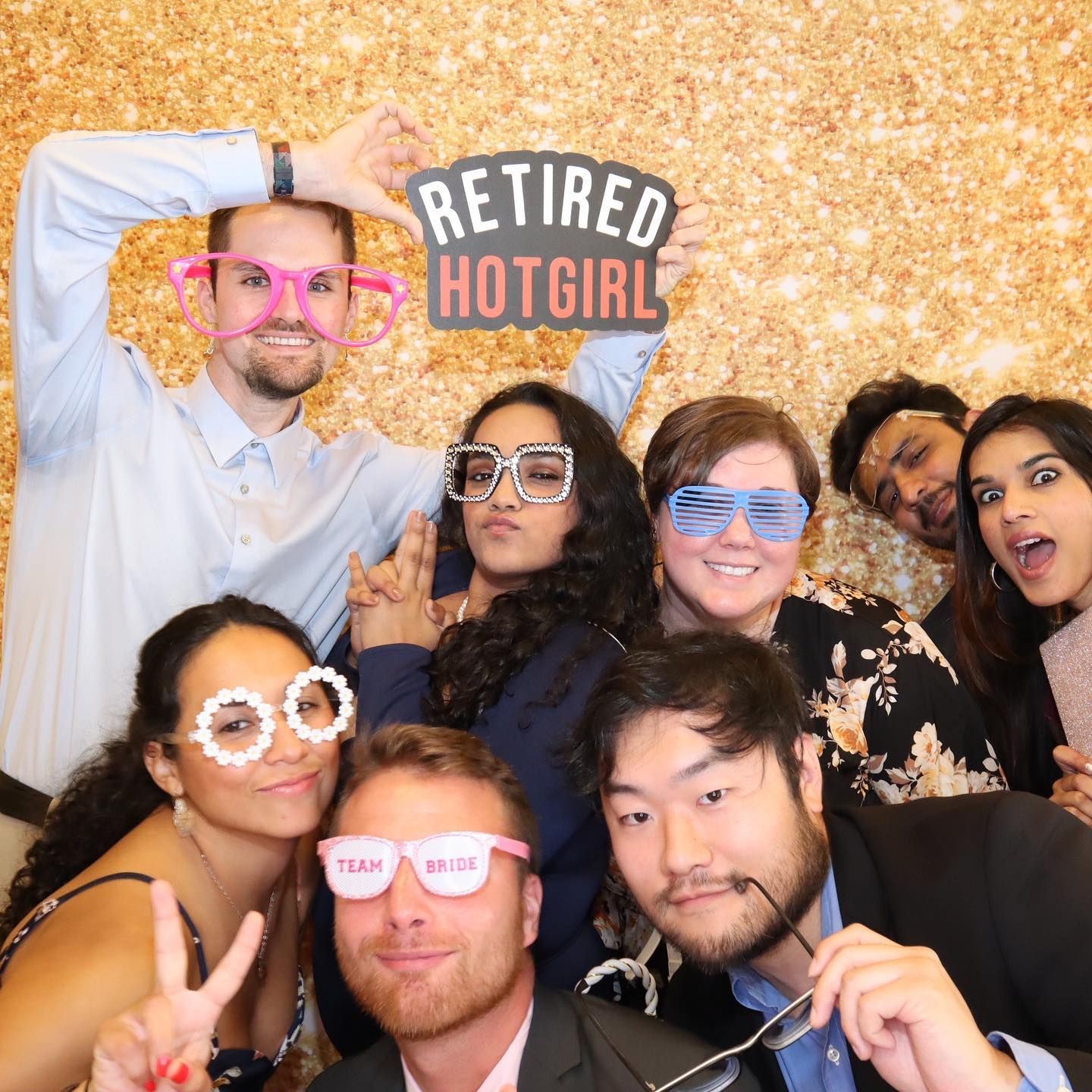Group of people in photo booth with props, smiling and posing with gold glitter background.