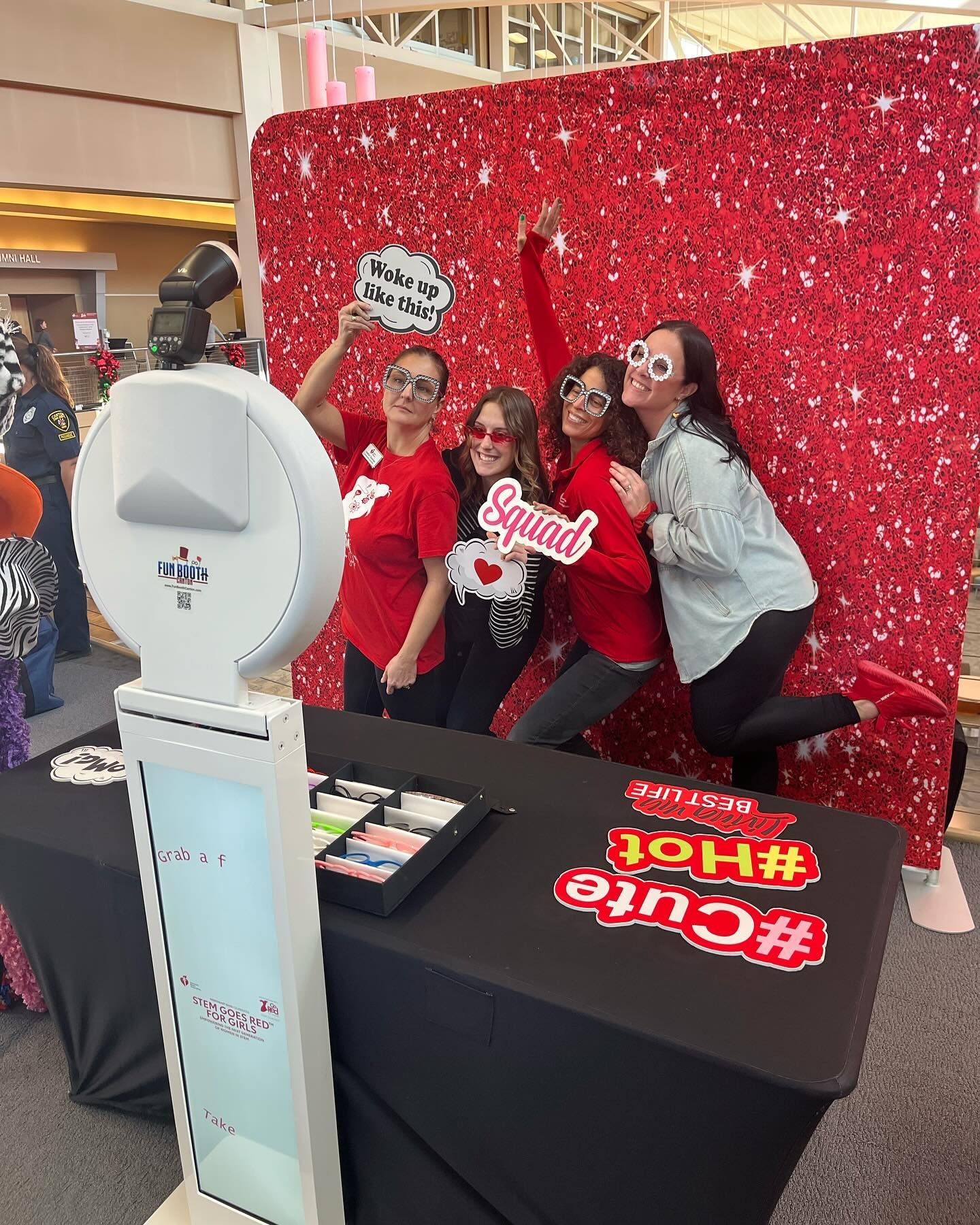 Four people pose with props in front of a red, sparkly backdrop near a photo booth.