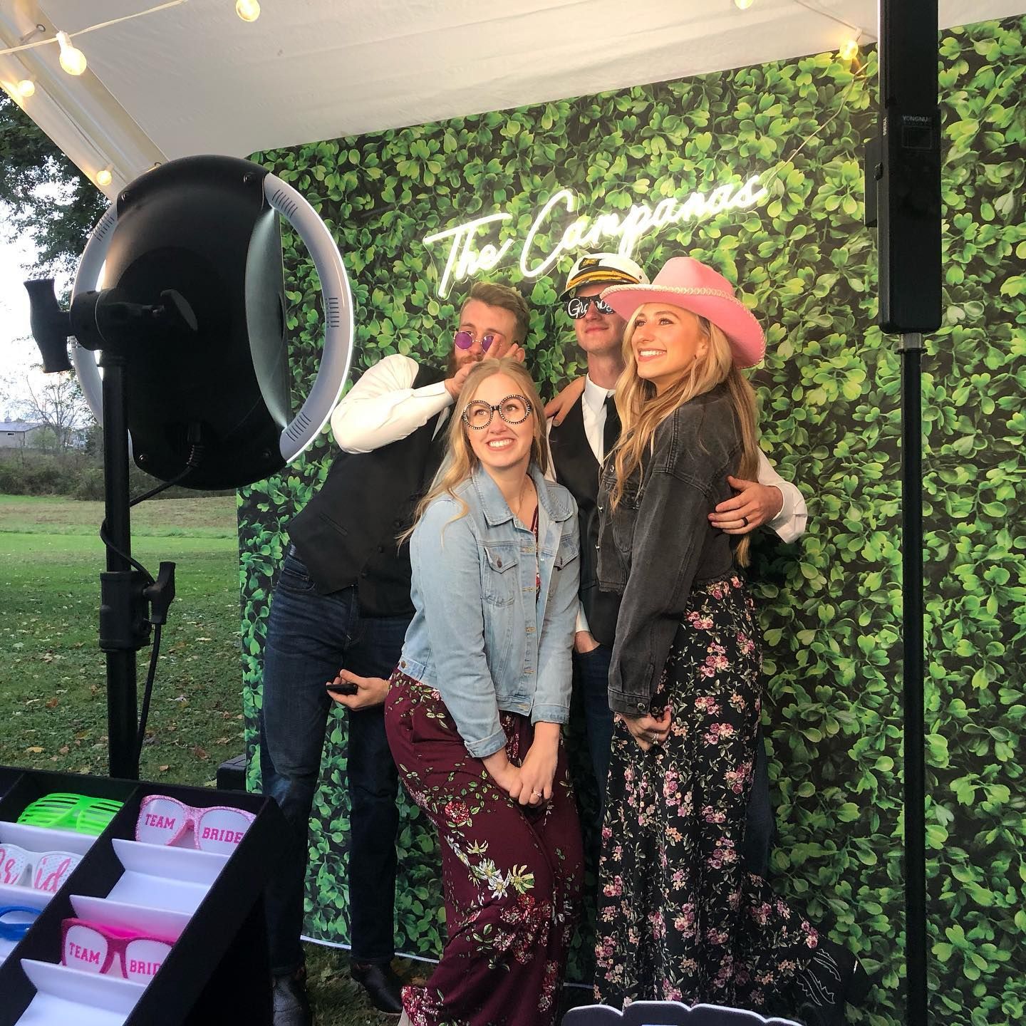 Four people pose at a photo booth with props. Green leafy backdrop and a neon sign.