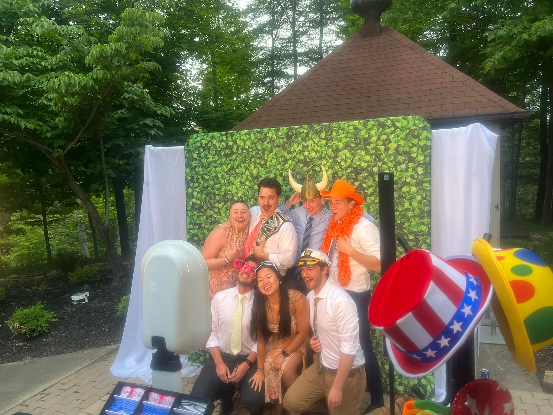 Group of people smiling, posing for a photo booth.  Props include hats, backdrop is a faux-hedge with a gazebo in background.