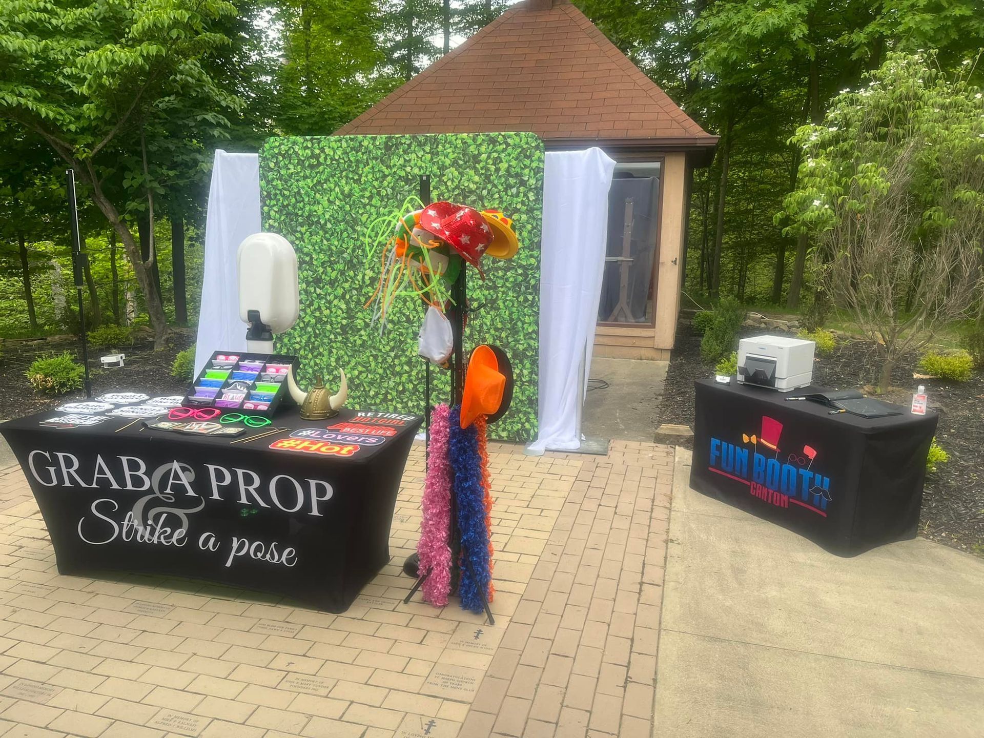 Photo booth setup outdoors; tables with props, backdrop, and equipment in front of a gazebo.