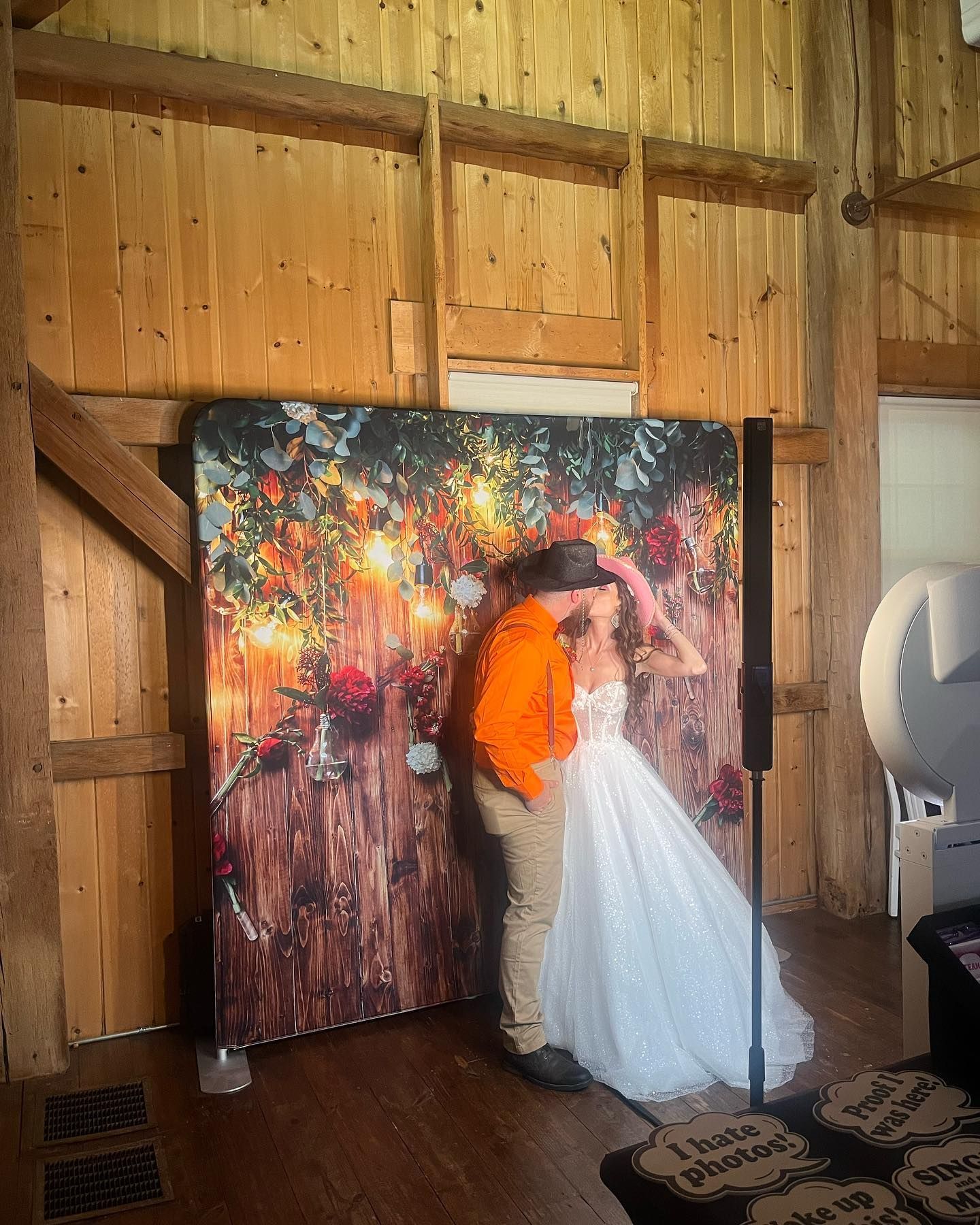 Couple kissing in front of a photo booth backdrop with floral and string light design in a wooden barn.