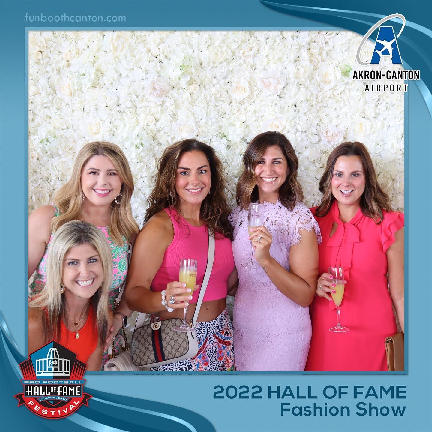 Five women at a fashion show pose with champagne. Floral wall backdrop.