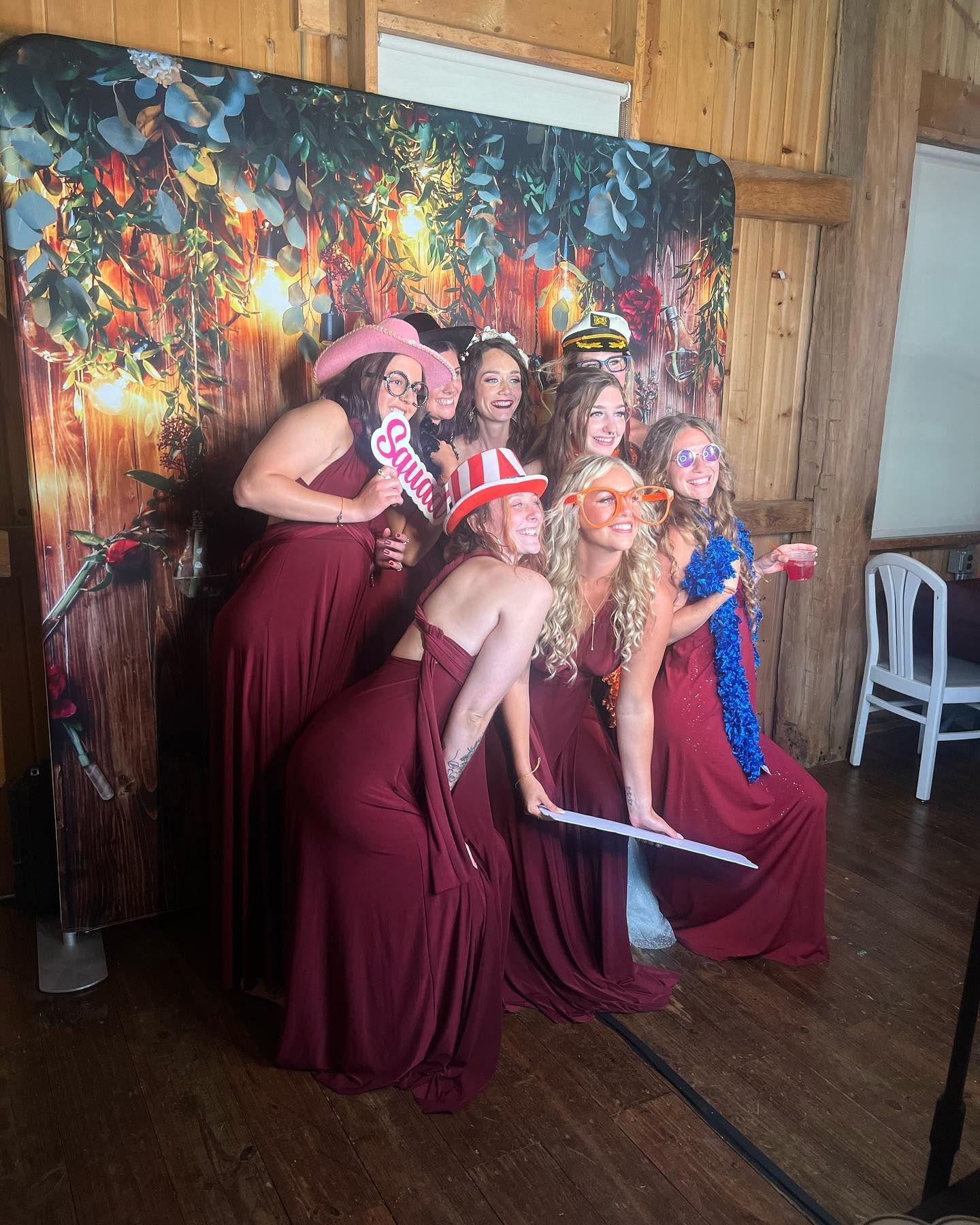 Bridesmaids pose in front of a photo backdrop; wearing maroon dresses, posing with props, and smiling.