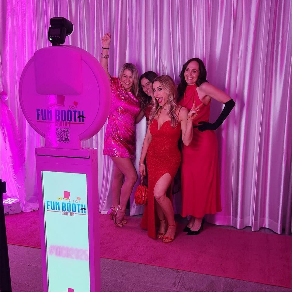Four women pose in front of a photo booth, on a pink carpet, lit by pink lights.