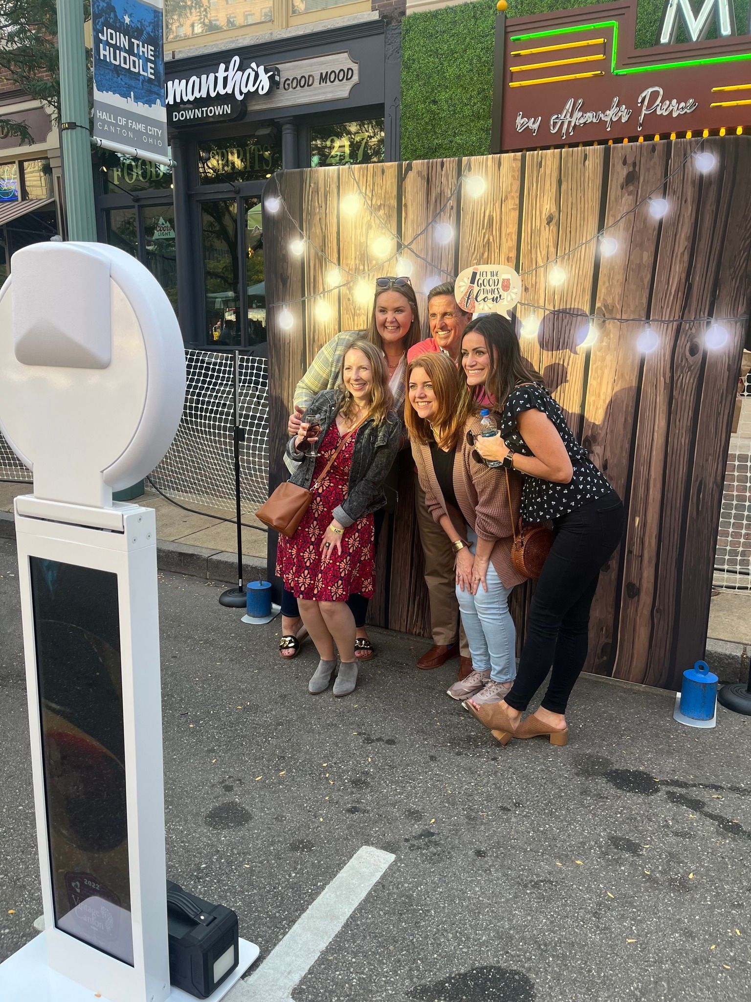 People posing for a photo booth with wooden backdrop and string lights on a city street.